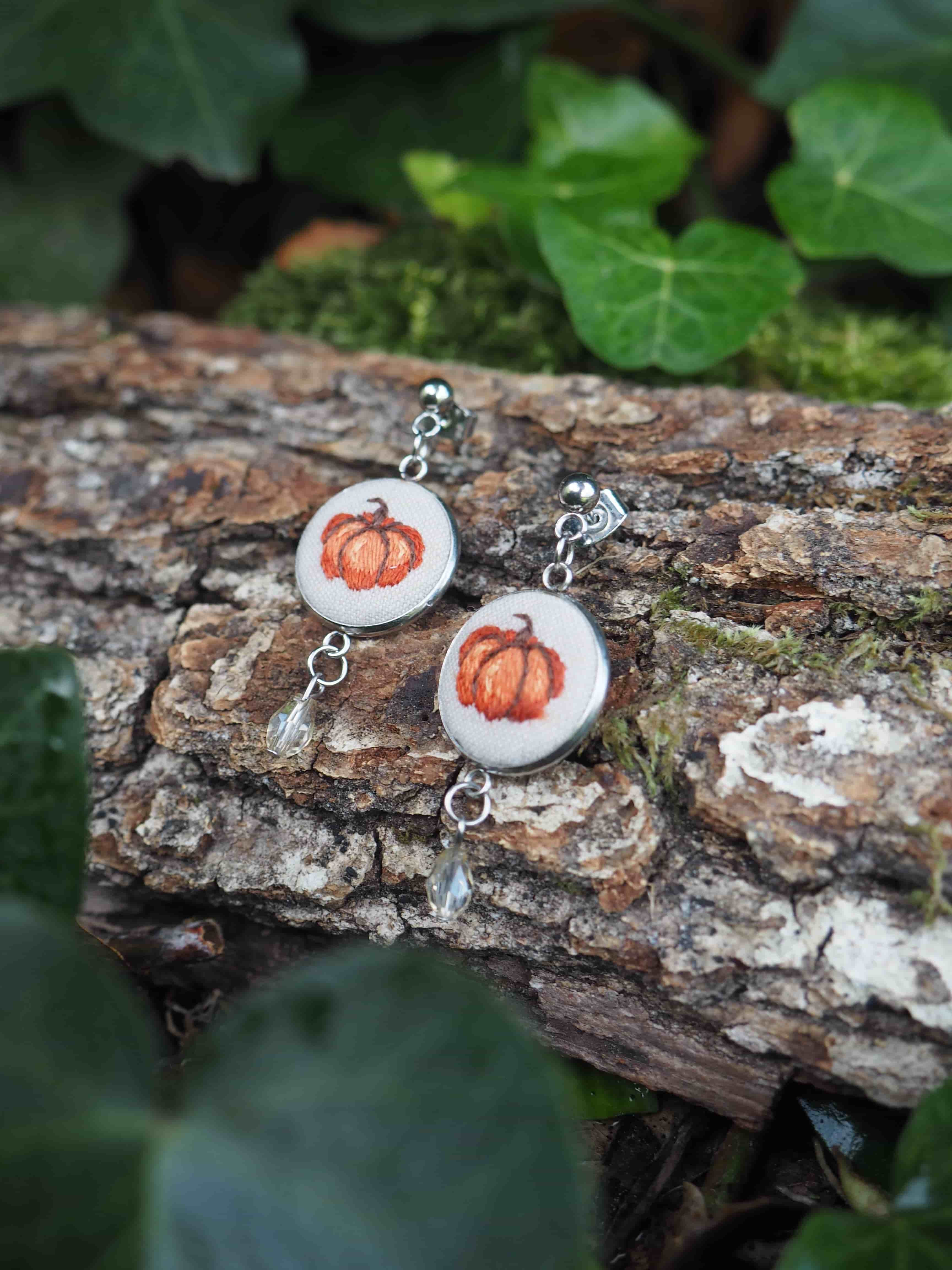 Cette image présente une paire de boucles d'oreilles artisanales disposées sur un tronc d'arbre dans un environnement naturel. Les bijoux sont composés de pendentifs circulaires blancs, probablement en céramique ou en résine, au centre desquels sont incrustées de petites citrouilles oranges miniatures, évoquant l'automne ou Halloween. Les boucles d'oreilles sont équipées de crochets métalliques argentés et reposent sur l'écorce rugueuse et texturée d'une branche ou d'un tronc. L'arrière-plan révèle un cadre forestier avec des feuilles vertes floues qui créent un joli contraste naturel avec les tons oranges des citrouilles. La photographie capture bien les détails des bijoux tout en les intégrant harmonieusement dans leur environnement naturel, créant une composition esthétique qui met en valeur le caractère artisanal et saisonnier de ces accessoires.