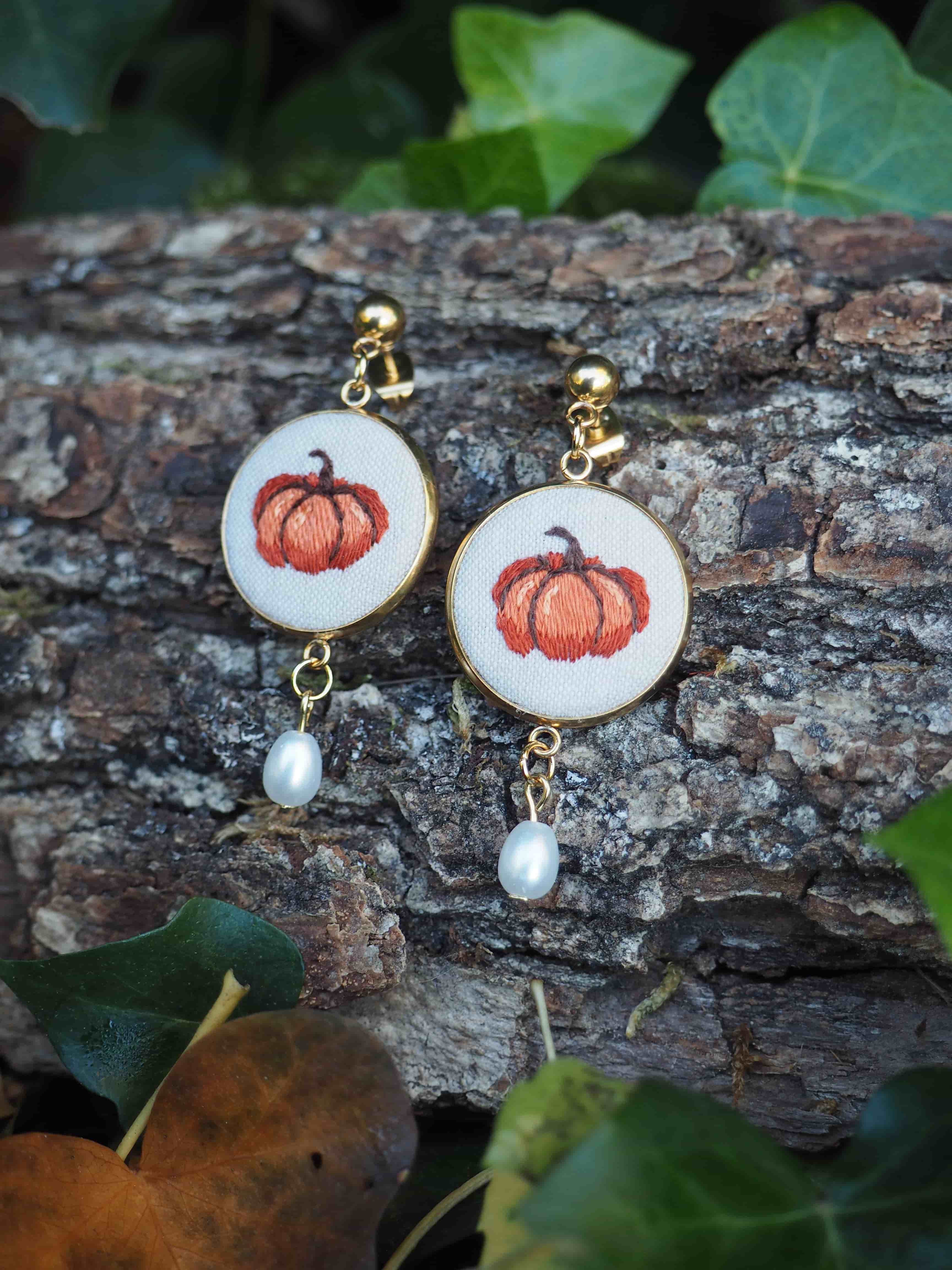 Cette image présente une paire de boucles d'oreilles artisanales disposées sur un tronc d'arbre dans un environnement naturel.  **Caractéristiques des bijoux :**
- Support principal : disques en bois clair ou matériau naturel de couleur beige/crème
- Motif brodé : citrouilles orange réalisées au fil, évoquant un thème automnal
- Finitions dorées : crochets et anneaux de connexion en métal doré
- Pendentifs : petites perles blanches nacrées suspendues sous chaque boucle **Mise en scène :**
- Posées sur l'écorce rugueuse et texturée d'un tronc d'arbre
- Environnement naturel avec des feuilles vertes visibles en arrière-plan
- Éclairage naturel qui met en valeur les détails de la broderie
- Composition harmonieuse entre les tons chauds des bijoux et les couleurs naturelles du décor Ces boucles d'oreilles semblent être des créations artisanales uniques, parfaites pour la saison automnale, alliant savoir-faire traditionnel de la broderie et design contemporain.