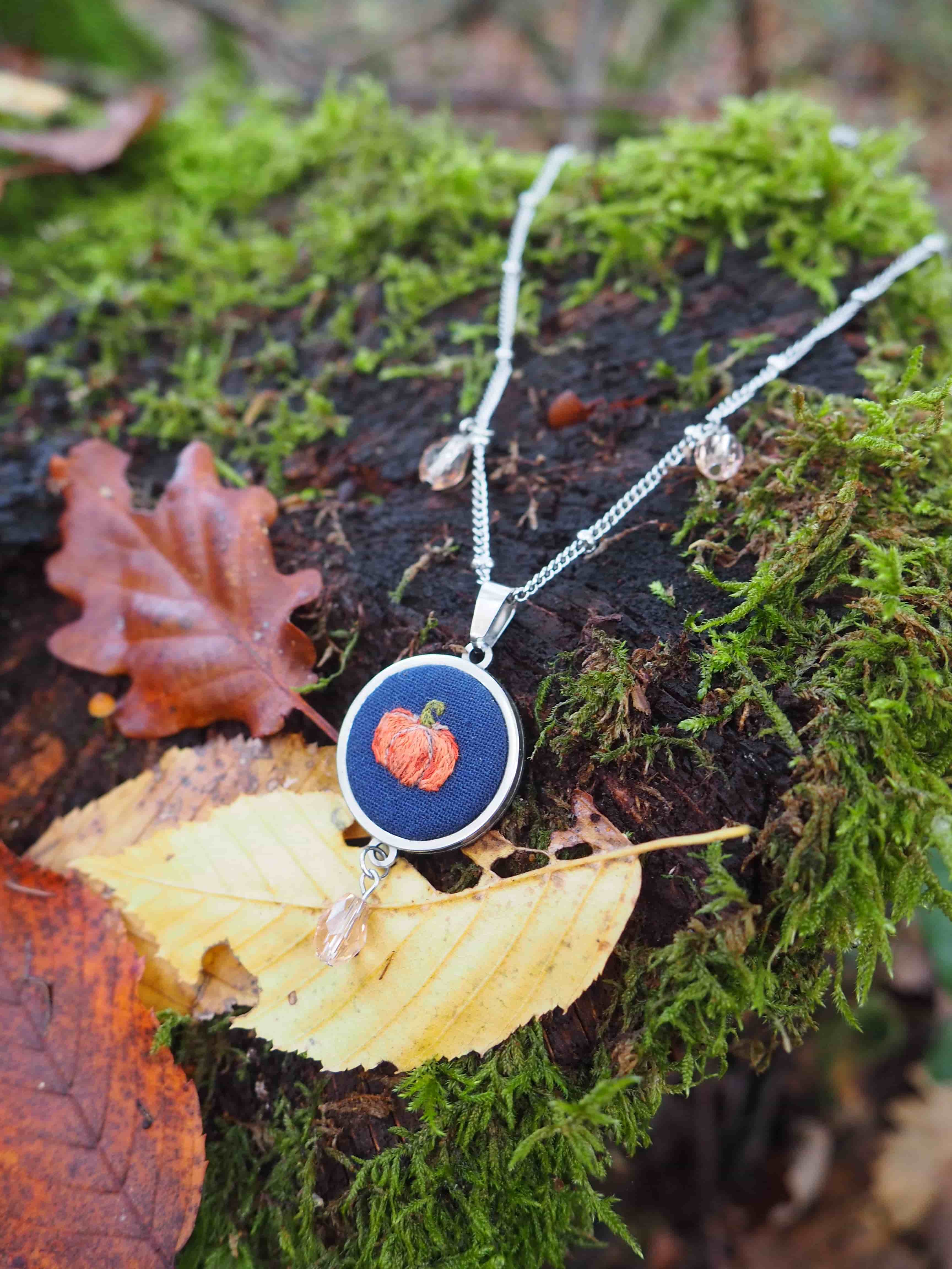Cette image présente un bijou artisanal photographié dans un cadre naturel très évocateur. Il s'agit d'un pendentif circulaire suspendu à une chaîne argentée, posé délicatement sur ce qui semble être un champignon ou un morceau de bois clair. Le pendentif lui-même est particulièrement intéressant : il présente un fond bleu profond sur lequel se détache un motif central de couleur rouge-orangé, possiblement une fleur ou un élément décoratif naturel. Le contraste entre ces couleurs chaudes et froides crée un effet visuel saisissant. L'environnement choisi pour cette mise en scène est remarquable : le bijou repose sur un lit de mousse verte luxuriante, parsemée de feuilles d'automne aux teintes cuivrées et rougeâtres. Cette composition évoque parfaitement l'atmosphère forestière et automnale, créant une harmonie parfaite entre l'objet artisanal et son environnement naturel. L'éclairage doux et la profondeur de champ utilisée mettent en valeur les détails du pendentif tout en créant un arrière-plan naturellement flou qui renforce l'aspect contemplatif de l'image.