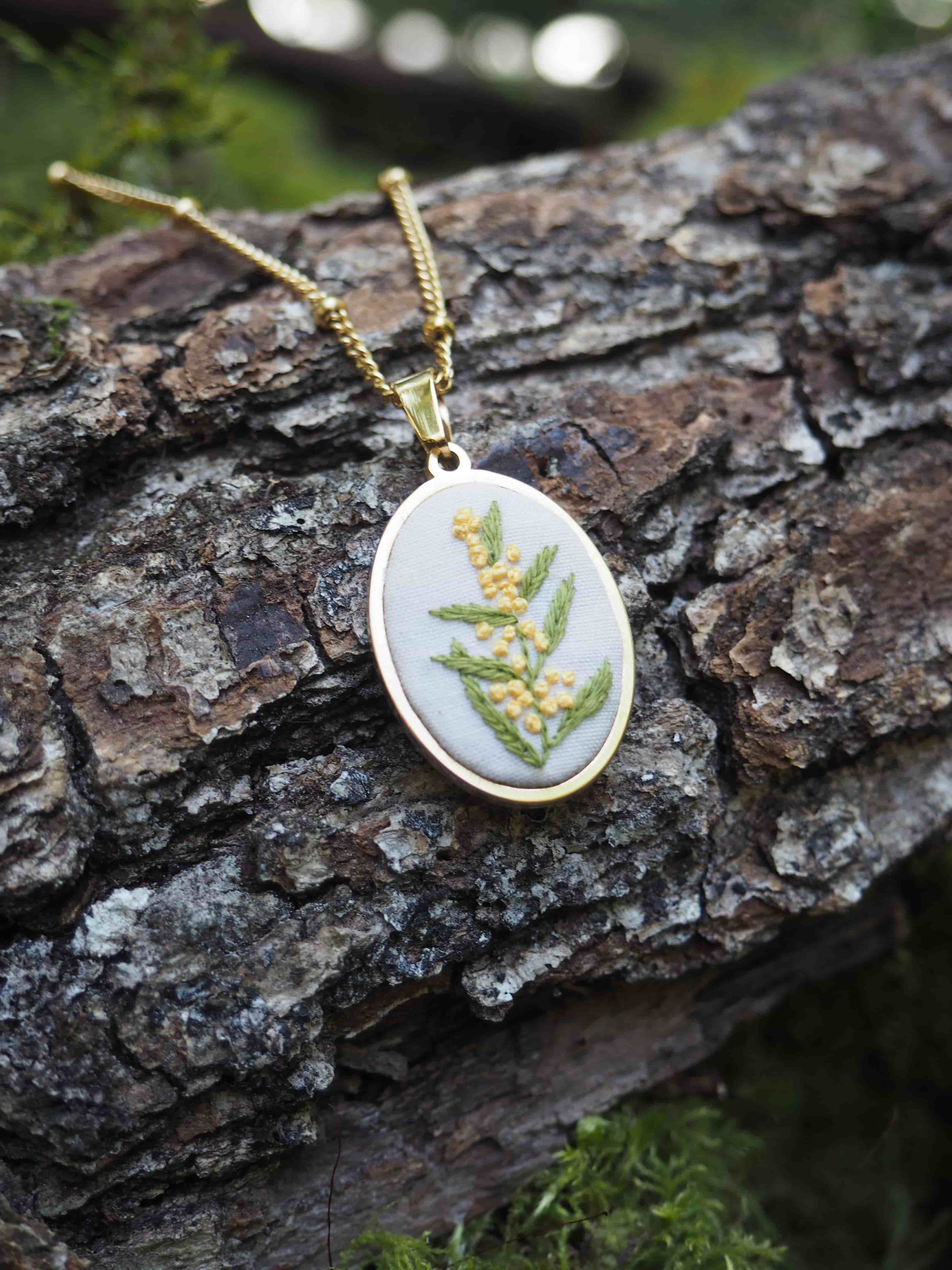 Cette image montre un délicat pendentif sur une chaîne dorée, photographié sur un morceau d'écorce d'arbre texturée. Le pendentif est de forme ovale ou ronde et présente un motif de fougère verte pressée ou peinte sur un fond blanc. C'est une pièce de bijouterie nature très élégante qui capture la beauté de la botanique. Le contraste entre le pendentif délicat et l'écorce rugueuse crée une belle composition, avec un peu de mousse verte visible dans le cadre, ajoutant à l'ambiance naturelle de la photo.
