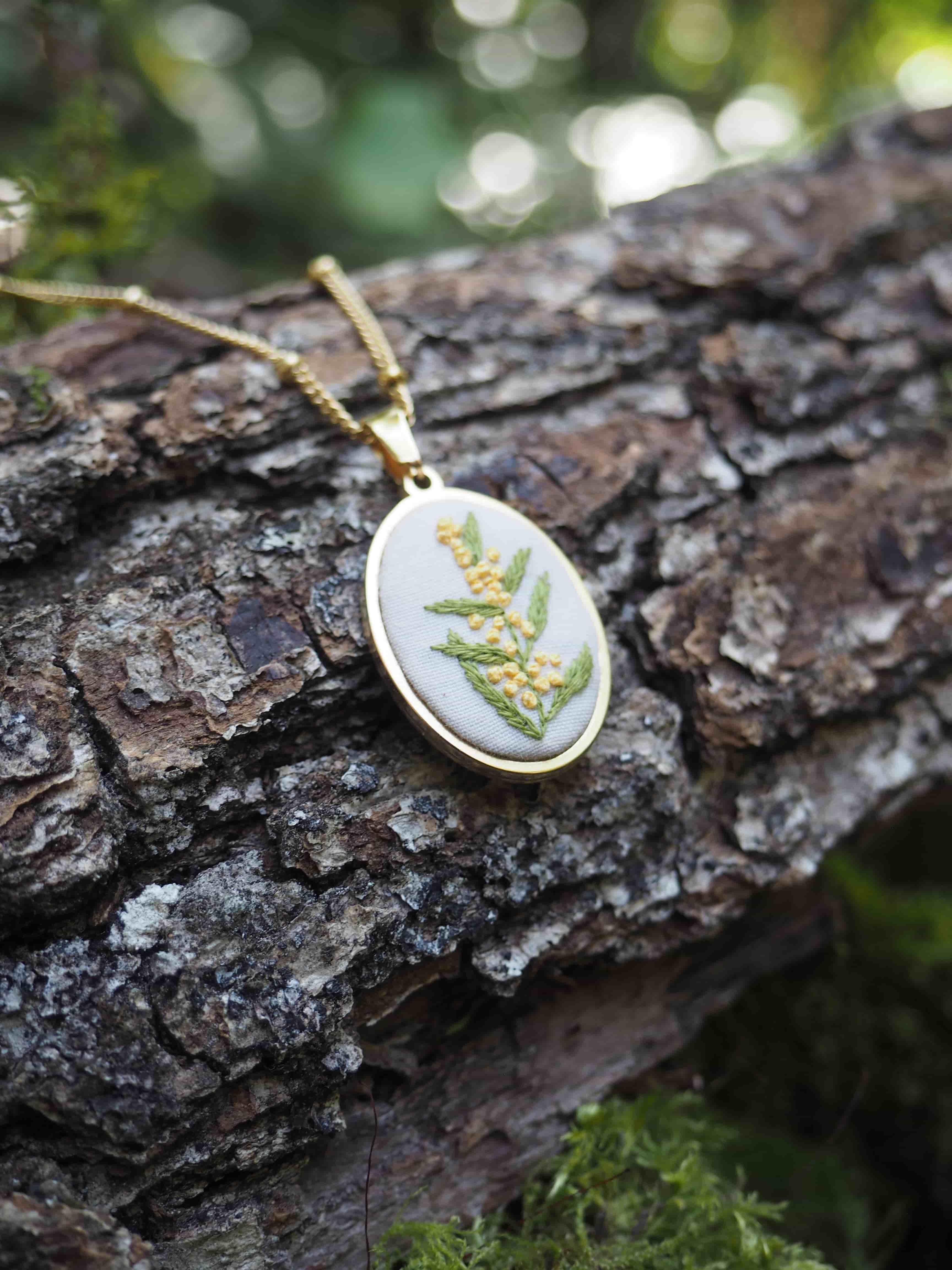 Cette image montre un délicat pendentif ovale posé sur un tronc d'arbre à l'écorce rugueuse. Le pendentif semble être fait d'argent ou d'un métal argenté, et contient de véritables feuilles ou fougères pressées et préservées sous verre ou résine. Il est suspendu à une chaîne dorée. L'arrière-plan est flou mais on peut distinguer des touches de vert et de la lumière naturelle créant un effet bokeh. C'est une belle composition qui met en valeur ce bijou artisanal inspiré par la nature, photographié dans un cadre naturel qui fait écho à ses motifs botaniques.