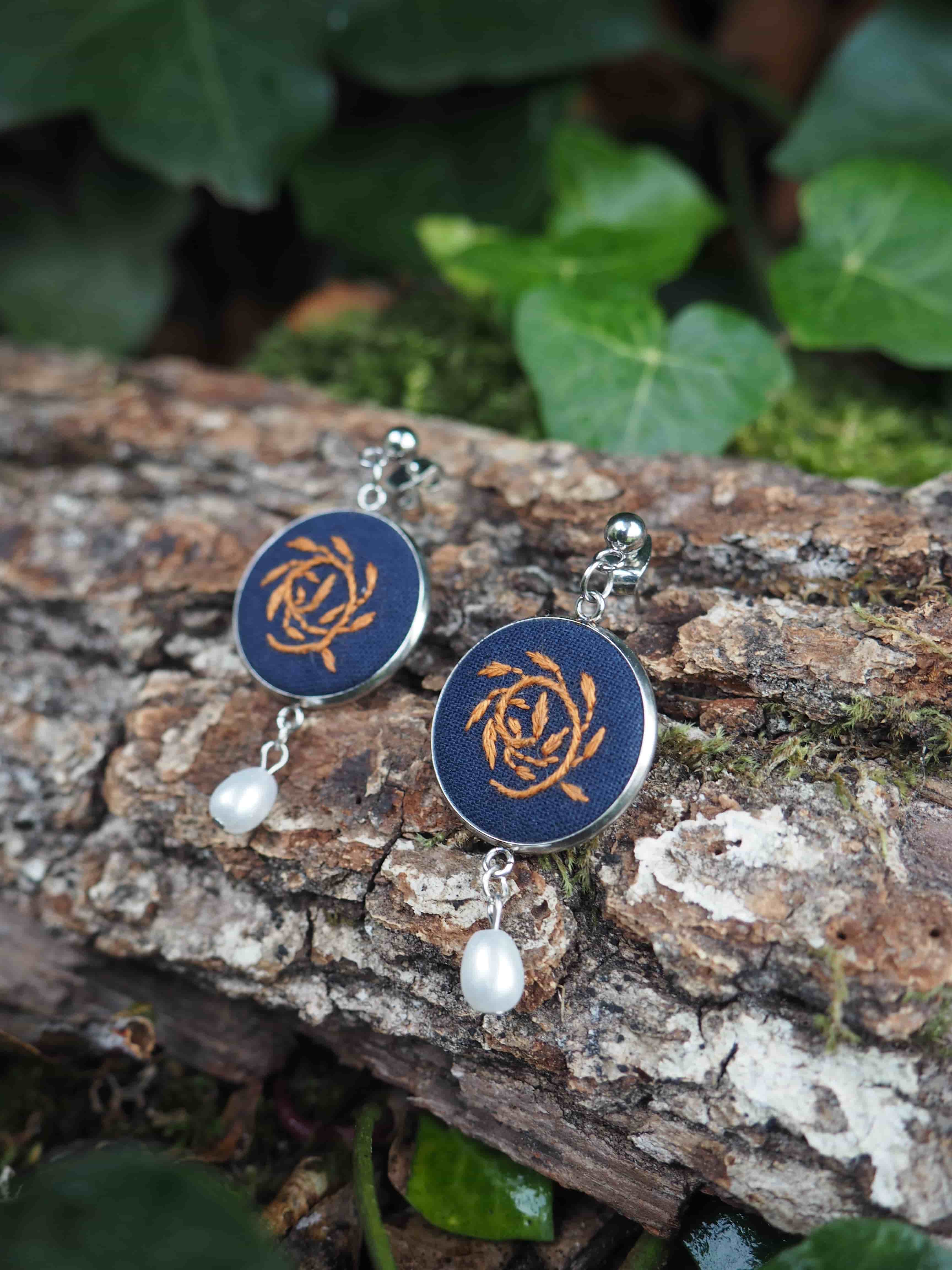 Cette image présente une paire de boucles d'oreilles artisanales disposées sur un tronc d'arbre dans un environnement naturel.  **Caractéristiques des bijoux :**
- Pendants circulaires de couleur bleu marine/indigo
- Motif décoratif doré au centre ressemblant à un nœud celtique ou à une rosace stylisée
- Petites perles blanches nacrées suspendues en dessous
- Crochets d'oreilles en métal argenté **Environnement :**
- Support : écorce rugueuse d'un tronc d'arbre avec des lichens blancs
- Arrière-plan : feuillage vert flou créant un bokeh naturel
- Éclairage doux et naturel qui met en valeur les couleurs des bijoux **Composition photographique :**
- Mise au point nette sur les boucles d'oreilles
- Profondeur de champ réduite pour isoler le sujet
- Contraste harmonieux entre les tons bleus des bijoux et les verts de la végétation Cette présentation met en valeur le caractère artisanal et naturel de ces bijoux, suggérant un style bohème ou ethnique.