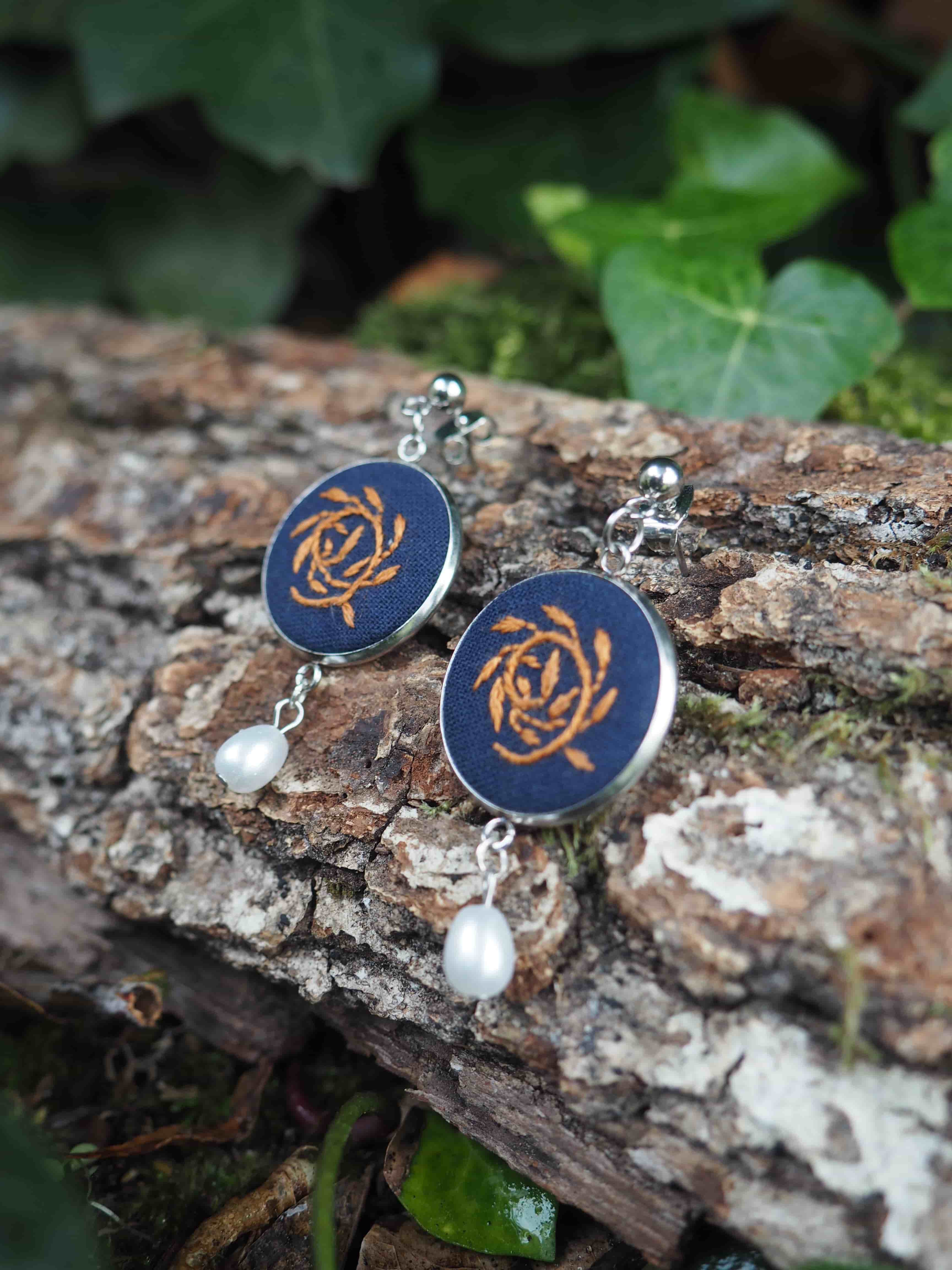 Cette image présente une paire de boucles d'oreilles artisanales posées sur un tronc d'arbre dans un environnement naturel.  **Caractéristiques des bijoux :**
- Pendants circulaires avec un fond bleu marine/noir
- Motif central doré représentant une rose stylisée ou un motif floral en spirale
- Perles blanches nacrées suspendues en bas de chaque boucle
- Crochets d'oreilles en métal argenté
- Style artisanal et élégant **Mise en scène :**
- Les boucles d'oreilles reposent sur l'écorce rugueuse et texturée d'une branche ou d'un tronc
- Arrière-plan naturel avec des feuilles vertes floues créant un effet de profondeur de champ
- Éclairage naturel doux qui met en valeur les détails et les couleurs
- Composition photographique soignée qui crée un contraste harmonieux entre l'objet manufacturé et l'environnement naturel L'ensemble évoque un style bohème ou artisanal, avec une esthétique qui marie l'élégance des bijoux et la beauté brute de la nature.