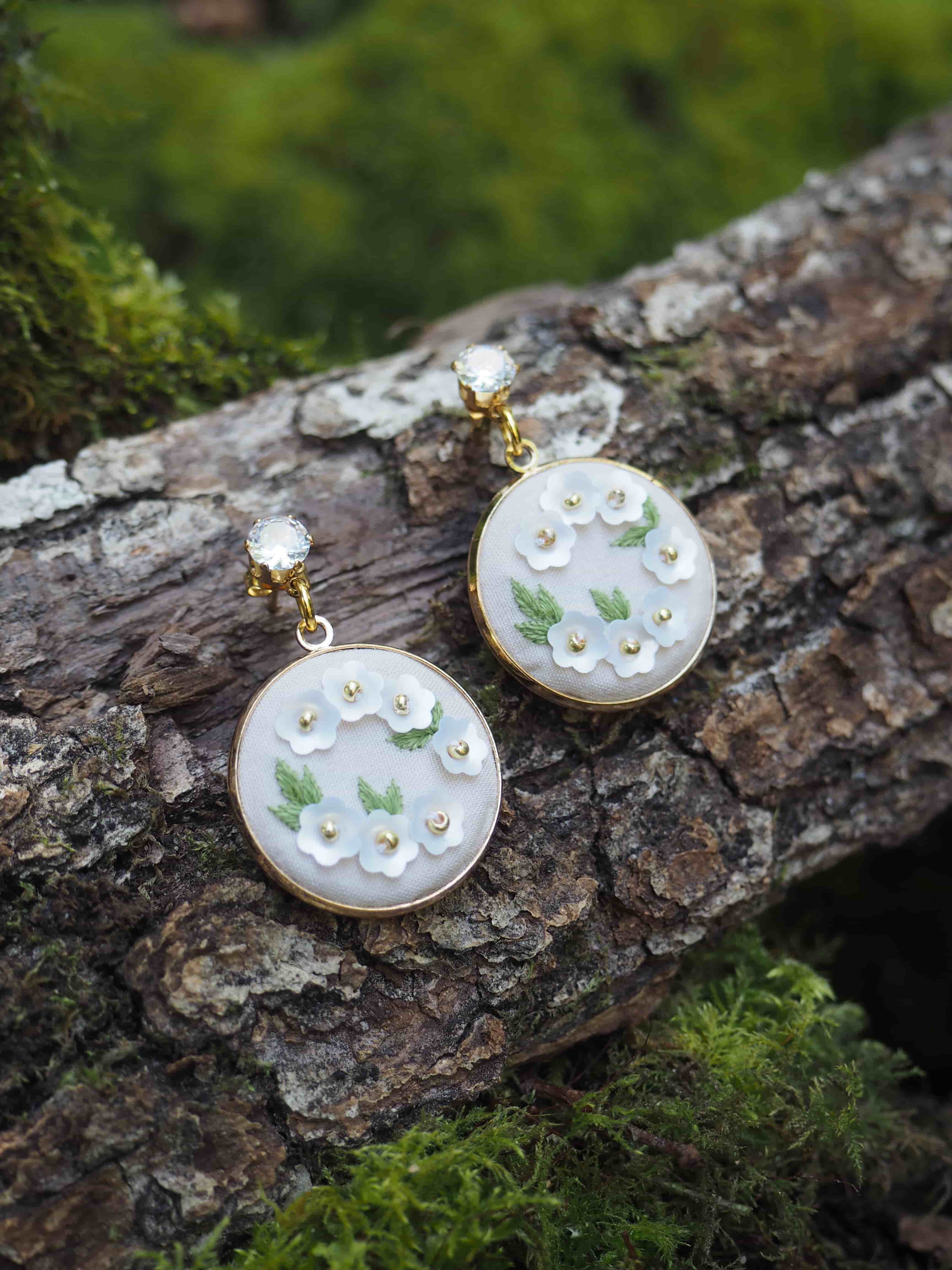Cette image montre une paire de boucles d'oreilles délicates posées sur un morceau de bois couvert de mousse. Les boucles d'oreilles sont de style vintage ou romantique, avec des médaillons ronds en nacre blanche. Chaque médaillon est décoré de petites fleurs blanches et de feuilles vertes en relief, ainsi que de petits strass ou perles qui ajoutent une touche scintillante. Les pendentifs sont montés sur des attaches dorées. La photo est prise dans un cadre naturel, avec le contraste entre la texture rugueuse du bois et la délicatesse des bijoux qui crée une composition très esthétique. Le fond vert flou et la mousse donnent une ambiance naturelle et féerique à l'image.