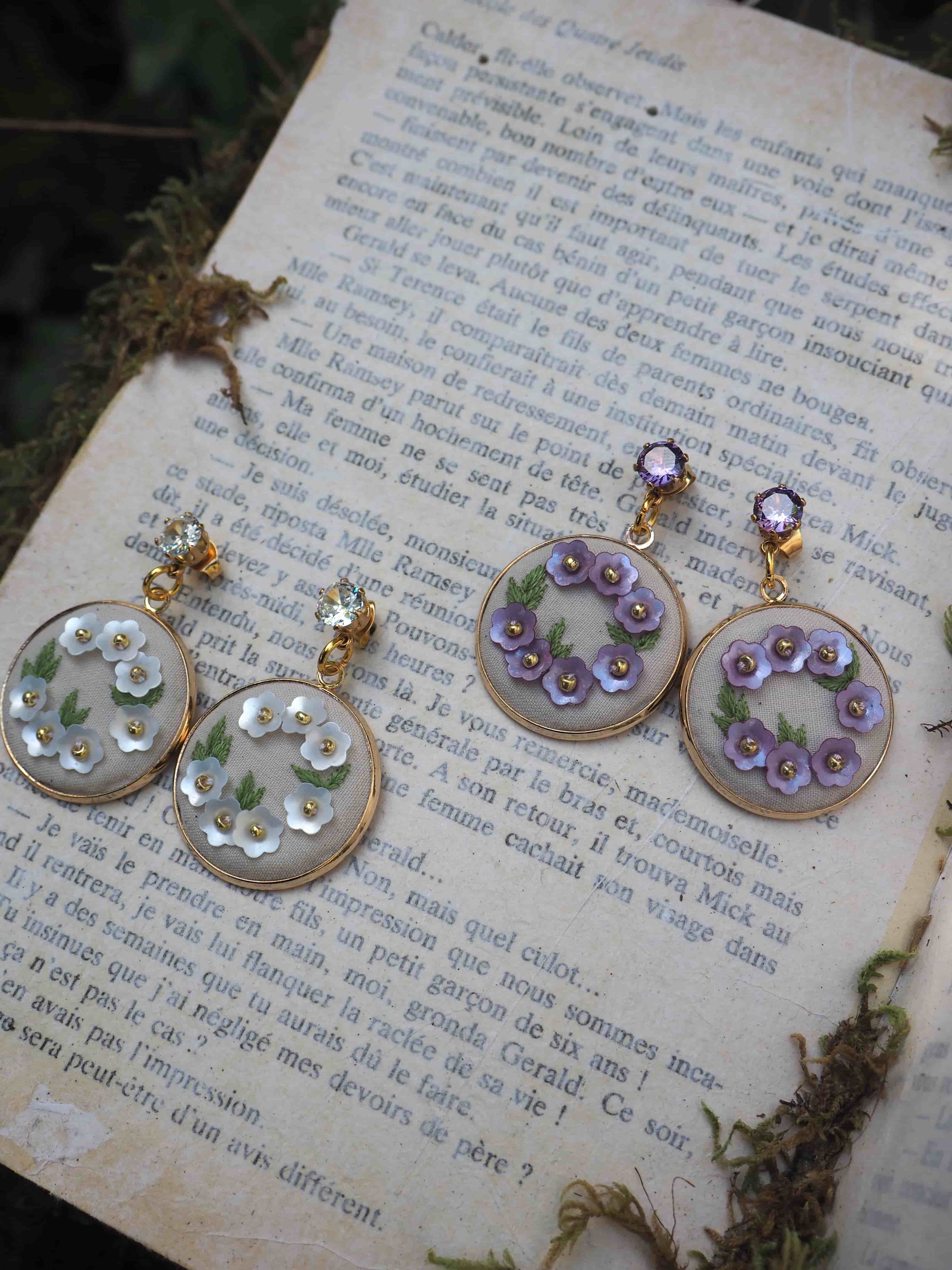 Ces belles boucles d'oreilles artisanales sont photographiées sur une page de livre ancien en français. Les bijoux semblent être composés de cercles transparents ornés de petites fleurs délicates - une paire avec des fleurs blanches/vertes et l'autre avec des fleurs violettes. Les cercles paraissent être en résine ou en verre, montés sur des supports dorés. Les fleurs miniatures donnent un aspect très romantique et vintage à ces bijoux. La mise en scène sur la page vieillie du livre ajoute une touche poétique et nostalgique à la photo.
