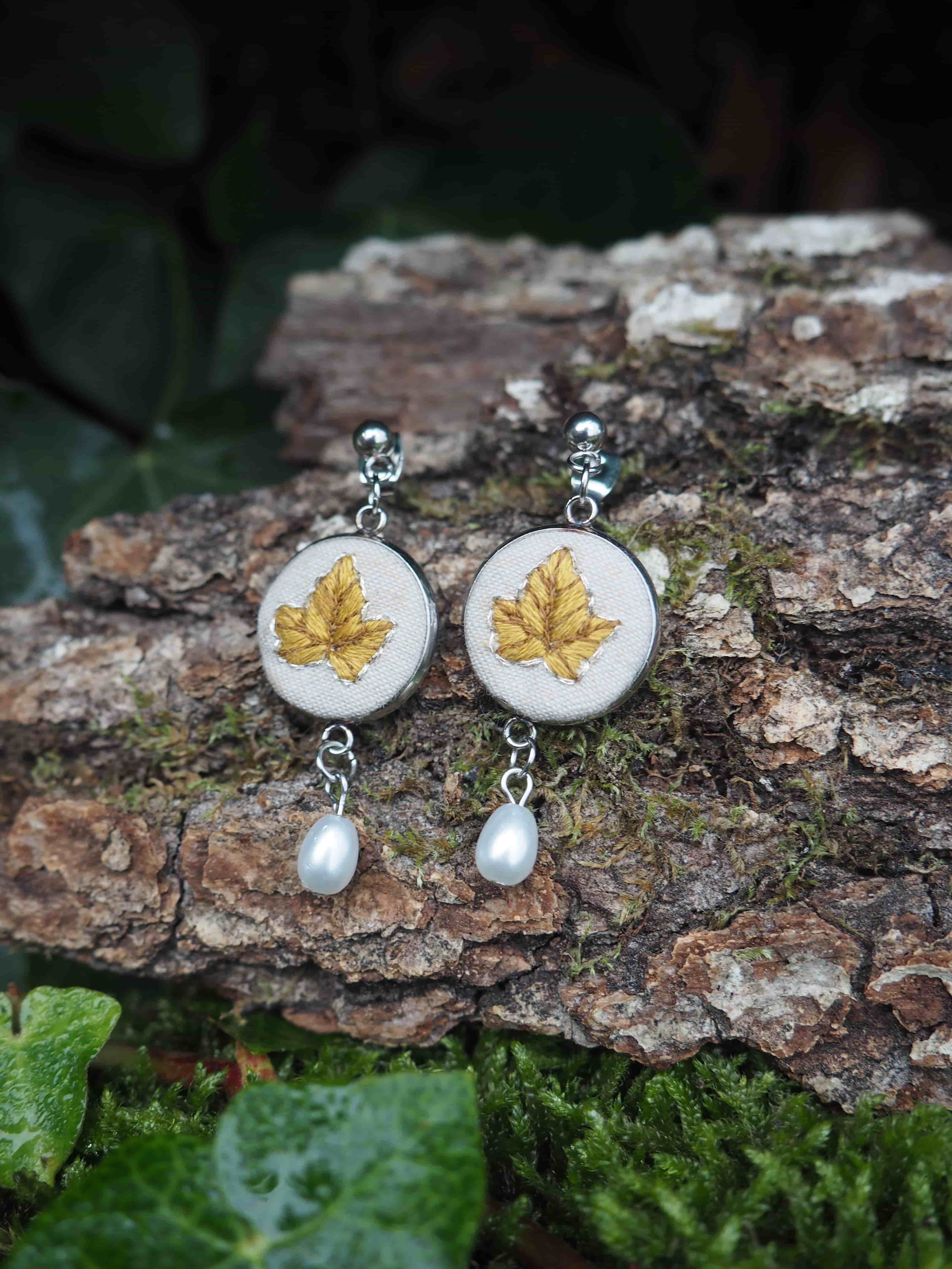 This image features a pair of elegant earrings resting on a piece of tree bark, surrounded by lush green foliage. The earrings are designed with a circular, silver-toned metal frame. Within each circle, there is a light-colored, possibly cream or off-white, fabric background on which a detailed golden-yellow leaf is embroidered. The leaf design appears to be a maple or ivy leaf, with visible veins and a slightly textured appearance from the embroidery. Suspended from the bottom of each circular earring is a single, lustrous white pearl, adding a touch of classic sophistication. The pearls are teardrop-shaped and reflect light softly. The background consists of a rough, textured piece of tree bark, covered in patches of vibrant green moss, suggesting a natural, outdoor setting. Below the bark, and slightly out of focus, are dark green leaves, possibly ivy, some of which appear to have water droplets on them, indicating a damp environment. The overall lighting is soft, highlighting the details of the earrings and the natural textures of the surroundings.