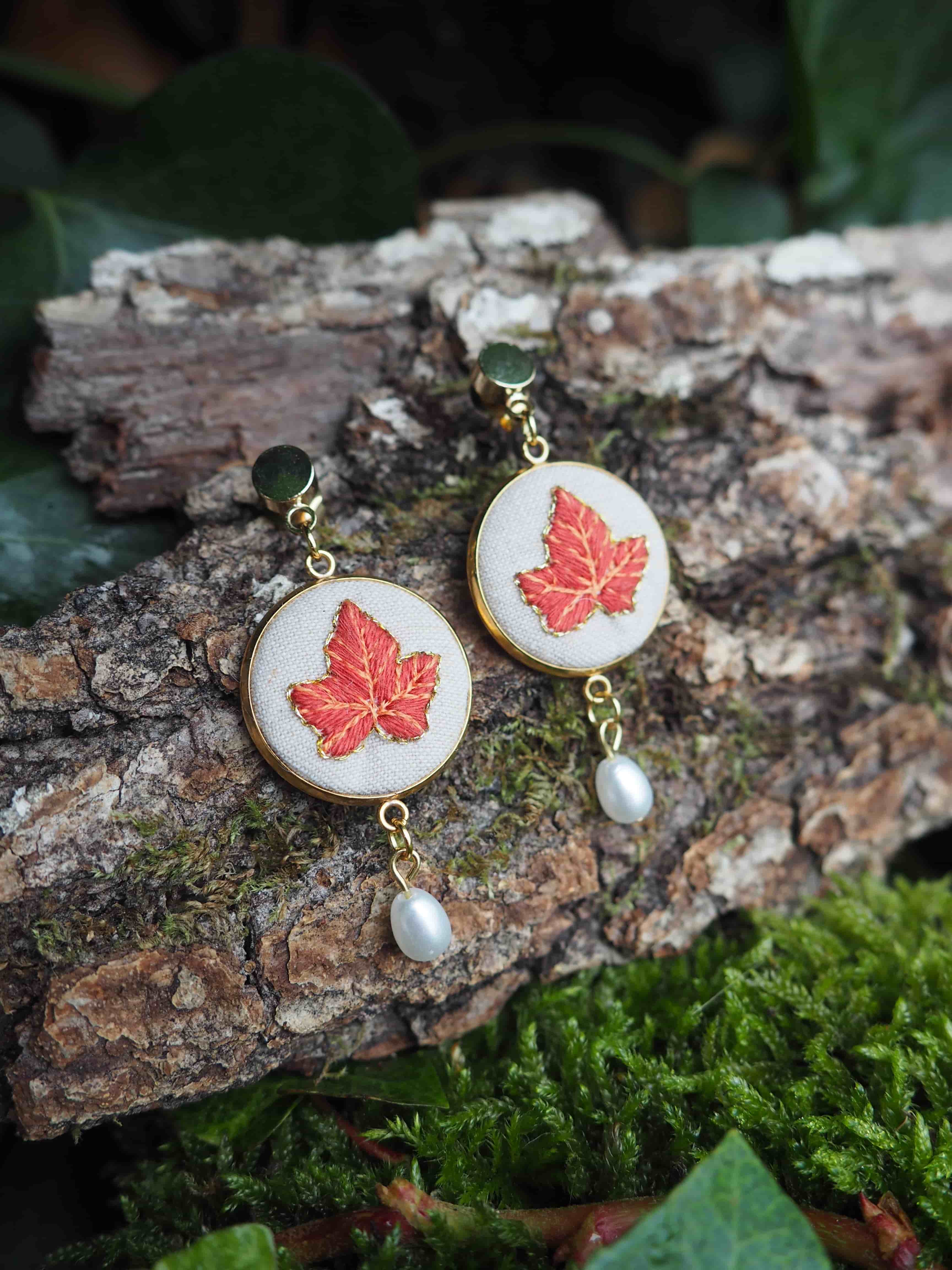 This close-up shot features a pair of elegant dangle earrings, each showcasing a beautifully embroidered red maple leaf on a circular, off-white fabric base. The embroidery is detailed, capturing the veins and shape of the leaf with vibrant red and subtle gold threads. The circular fabric is encased in a thin, golden metal frame, which also forms the stud and the dangle components of the earrings. Each earring is adorned with a small, lustrous white pearl that dangles gracefully from the bottom of the circular design, adding a touch of sophistication. The golden metal components appear polished and reflective. The earrings are artfully displayed on a piece of rough, textured tree bark, which is partially covered in patches of vibrant green moss. In the background, out of focus, are hints of dark green foliage, suggesting a natural, outdoor setting. The lighting is soft, highlighting the intricate details of the earrings and creating a gentle contrast with the rustic backdrop.