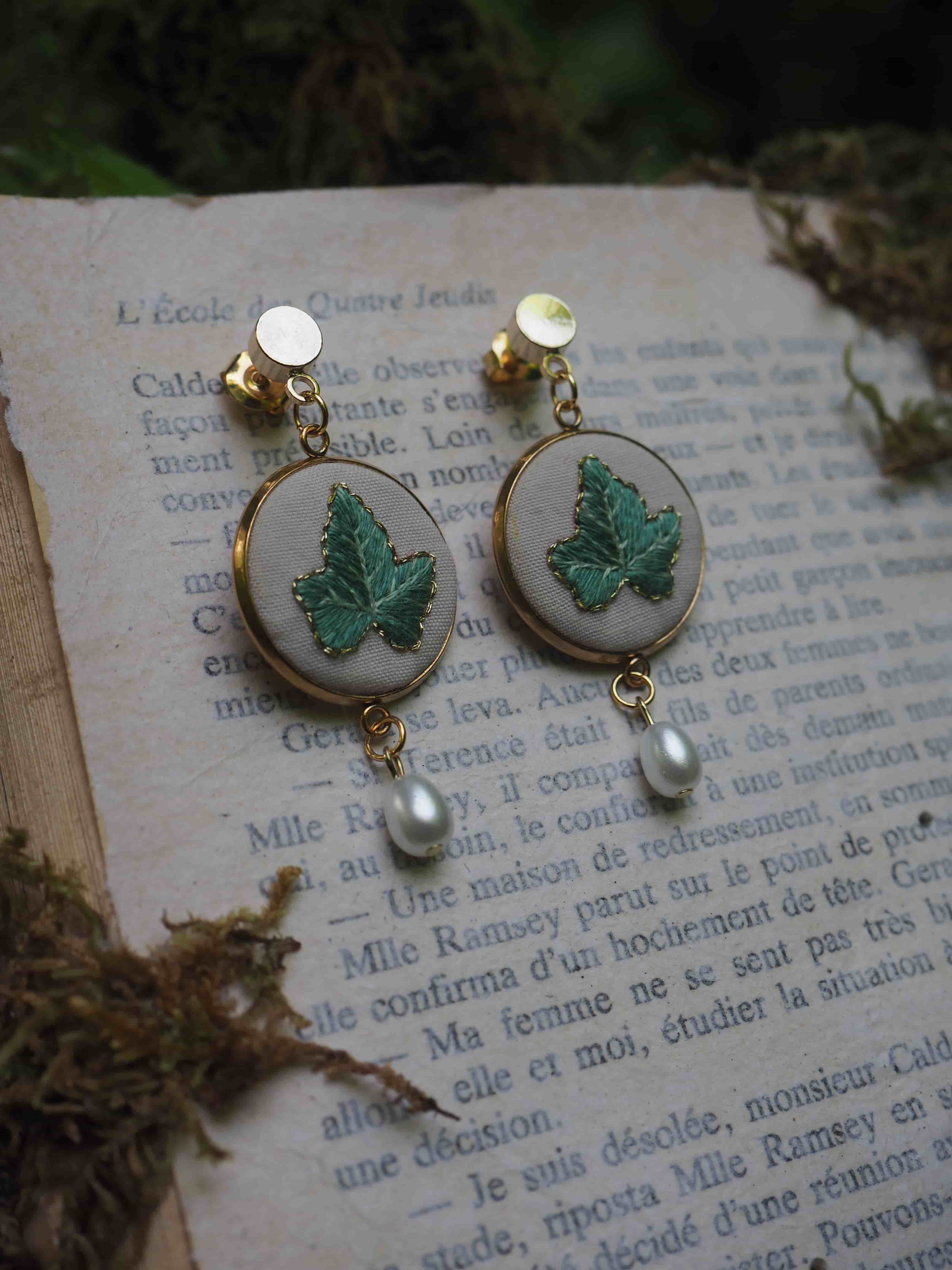 Cette image montre une paire de boucles d'oreilles élégantes posées sur une page de livre en français. Les boucles d'oreilles sont composées de plusieurs éléments : 1. Des clips ou clous dorés en haut
2. Un médaillon rond avec un fond clair (probablement beige ou blanc)
3. Une feuille verte brodée ou peinte sur chaque médaillon
4. Une petite perle blanche suspendue au bas de chaque boucle d'oreille Le style est très romantique et vintage, avec un aspect fait main ou artisanal. Les boucles d'oreilles sont photographiées sur ce qui semble être une page d'un livre ancien en français, créant une ambiance nostalgique et poétique. L'ensemble est très délicat et féminin, parfait pour un style bohème ou romantique.