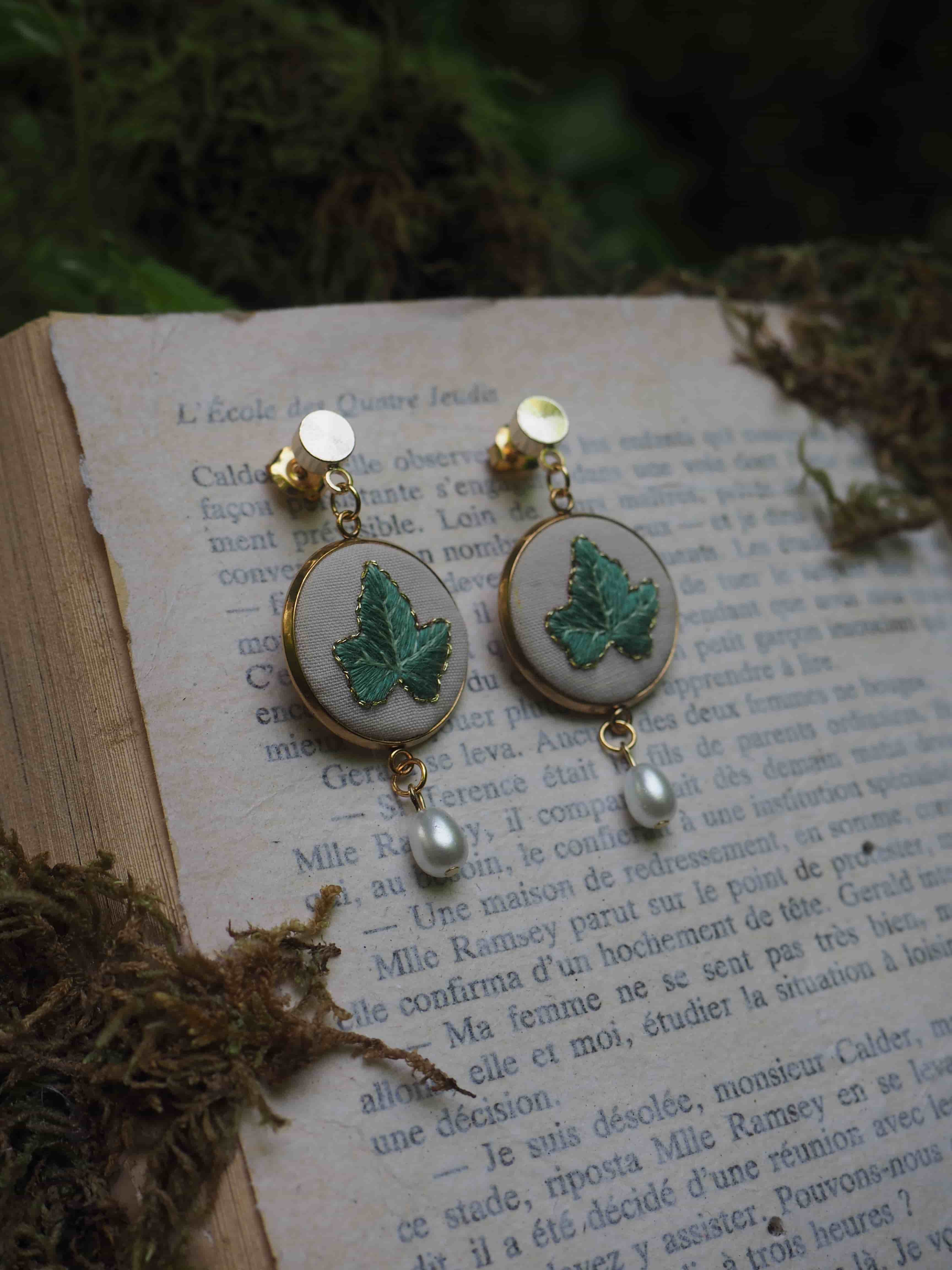 Cette image montre une paire de boucles d'oreilles élégantes posées sur une page de livre en français. Les boucles d'oreilles sont composées de médaillons ronds blancs ornés de papillons verts brodés ou peints. Au bas de chaque boucle d'oreille pend une petite perle nacrée. Les boucles d'oreilles semblent être de style vintage ou artisanal. L'arrière-plan montre du texte français imprimé sur une page, créant une ambiance romantique et littéraire. La photo a une atmosphère douce et poétique, avec une légère présence de verdure floue en arrière-plan.