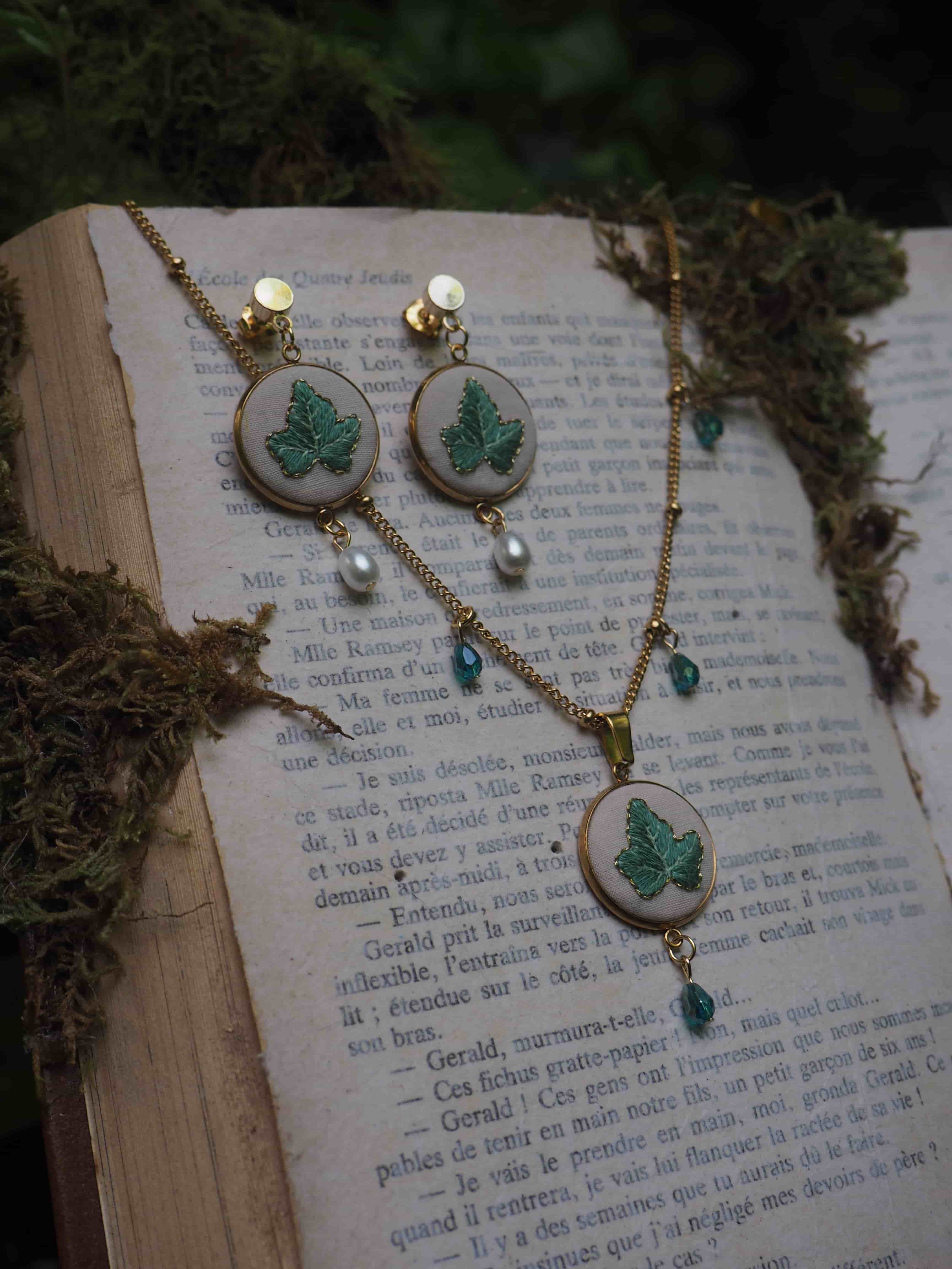 Cette image montre un ensemble de bijoux délicat posé sur un livre ouvert. L'ensemble se compose d'une paire de boucles d'oreilles et d'un collier assortis. Chaque pièce présente une broderie verte en forme de feuille ou de trèfle sur un fond blanc, encadrée dans ce qui semble être des cercles en métal doré. Les bijoux sont ornés de petites perles et de cristaux bleu-vert qui pendent en dessous des motifs brodés. L'arrière-plan montre les pages d'un livre en français avec du texte imprimé, et il y a ce qui semble être de la mousse ou des éléments naturels séchés disposés autour des bijoux, créant une ambiance naturelle et romantique. L'ensemble a une esthétique très délicate et vintage, avec une belle harmonie entre les éléments naturels et les bijoux artisanaux.