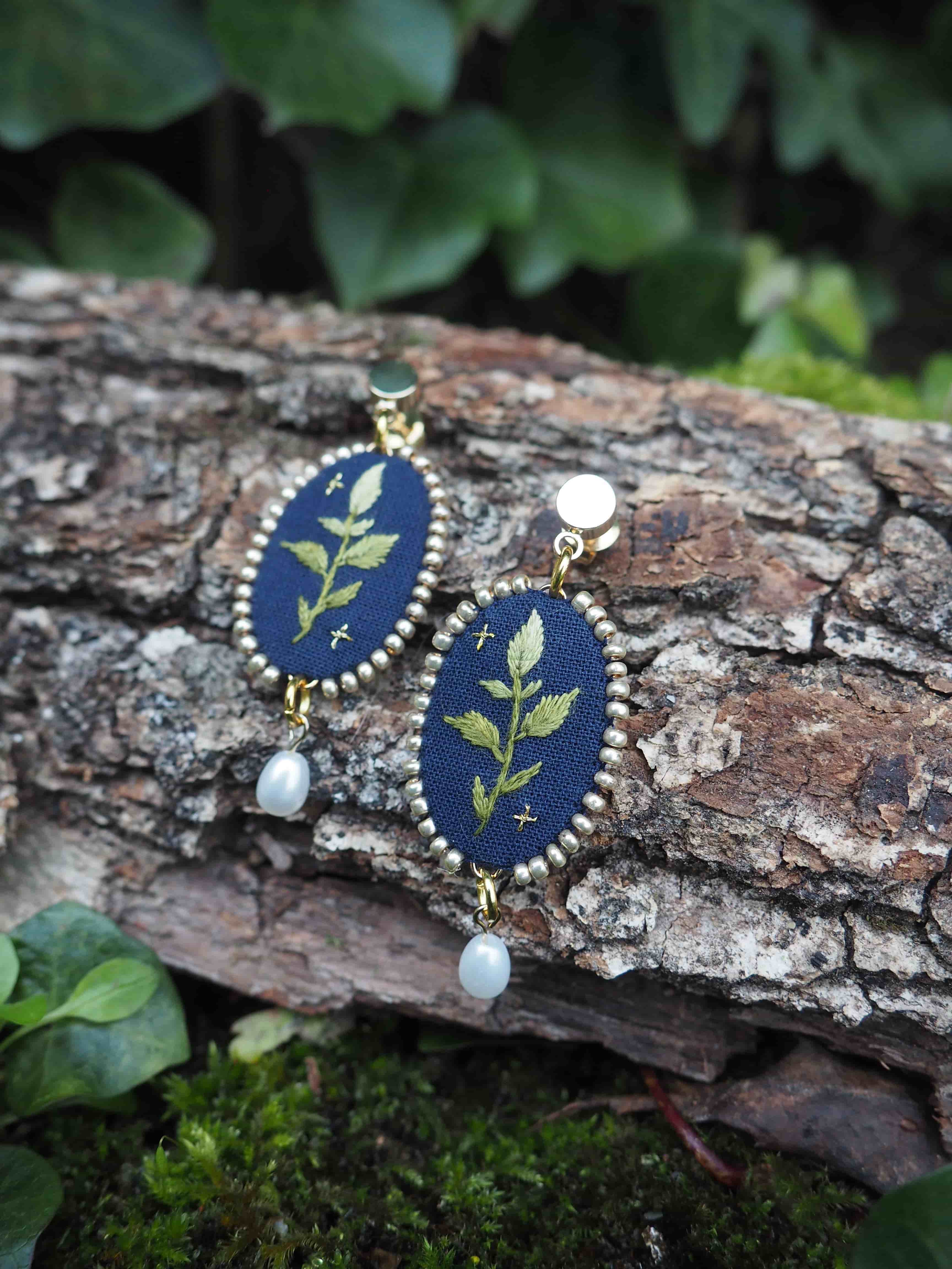 L'image montre une paire de boucles d'oreilles artisanales posées sur un morceau d'écorce d'arbre. Les boucles d'oreilles sont ovales avec un fond bleu foncé sur lequel est brodé un motif de feuille ou de branche en vert clair. Le contour des ovales semble être en métal doré, donnant un aspect élégant à l'ensemble. Chaque boucle d'oreille est ornée d'une petite perle blanche suspendue en bas, ajoutant une touche délicate. Le contraste entre le bleu profond du fond et le vert tendre de la broderie crée un effet visuel très attrayant. L'arrière-plan montre l'écorce rugueuse et texturée sur laquelle les boucles d'oreilles sont posées, ainsi que quelques feuilles vertes floues en arrière-plan, suggérant un cadre naturel en extérieur. Cette mise en scène met en valeur l'aspect artisanal et naturel des bijoux. Ces boucles d'oreilles semblent être un bel exemple d'artisanat mêlant broderie et joaillerie, avec une inspiration clairement tirée de la nature.