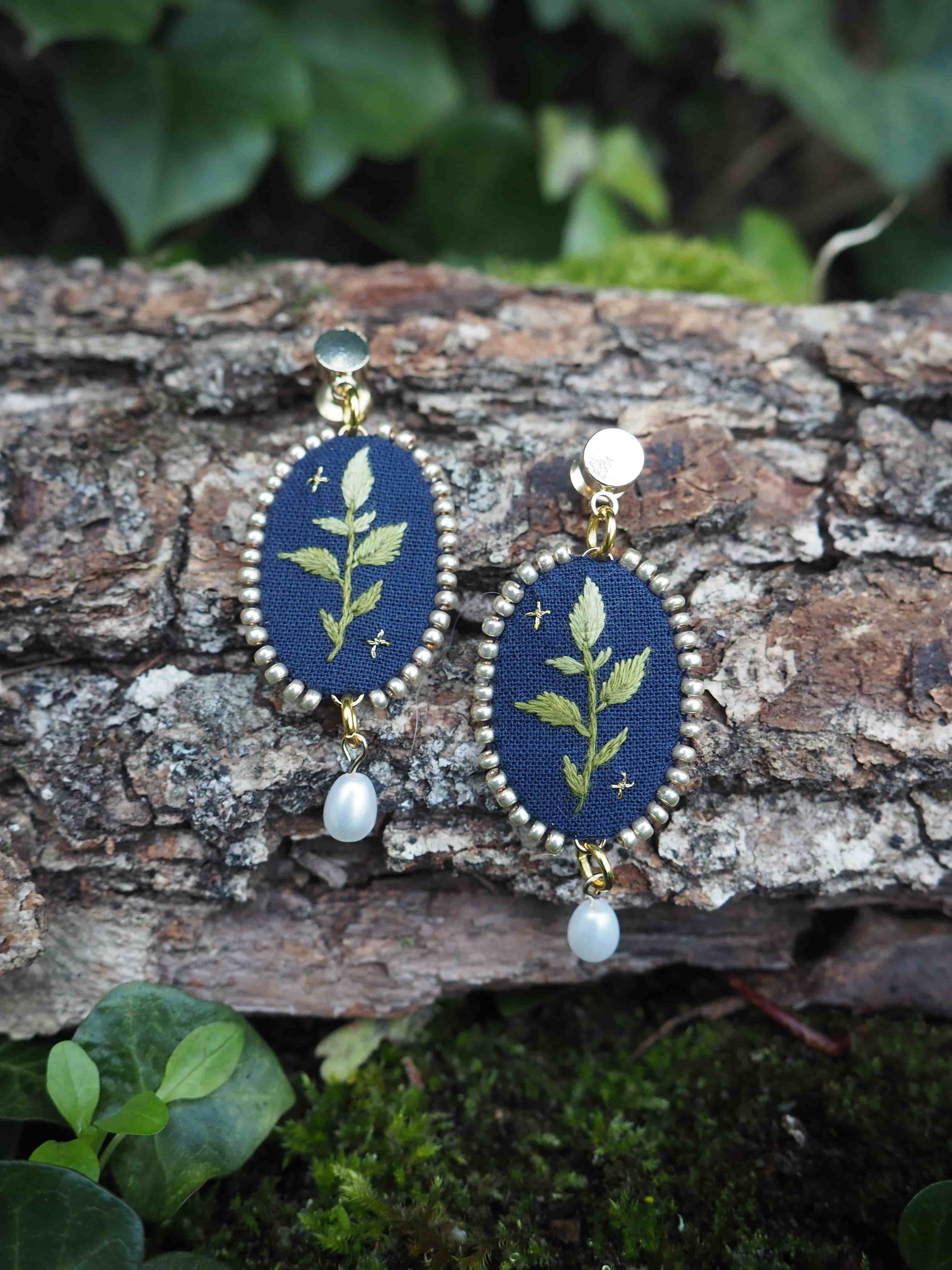 L'image montre une paire de boucles d'oreilles délicates et artisanales posées sur un morceau d'écorce d'arbre. Les boucles d'oreilles ont une forme ovale et sont composées d'un fond bleu foncé sur lequel est brodé un motif de feuille ou de branche verte. Elles sont entourées d'un cadre doré et ornées d'une petite perle blanche suspendue en bas. Le cadre naturel de l'image met en valeur ces bijoux faits main. L'écorce rugueuse et texturée contraste avec la finesse des boucles d'oreilles. En arrière-plan et autour, on peut voir de la verdure floue, probablement des feuilles, ce qui donne une ambiance naturelle et forestière à la scène. Cette photographie semble mettre en avant l'artisanat et la beauté des bijoux inspirés par la nature, en les plaçant dans un environnement qui reflète leur thème botanique.