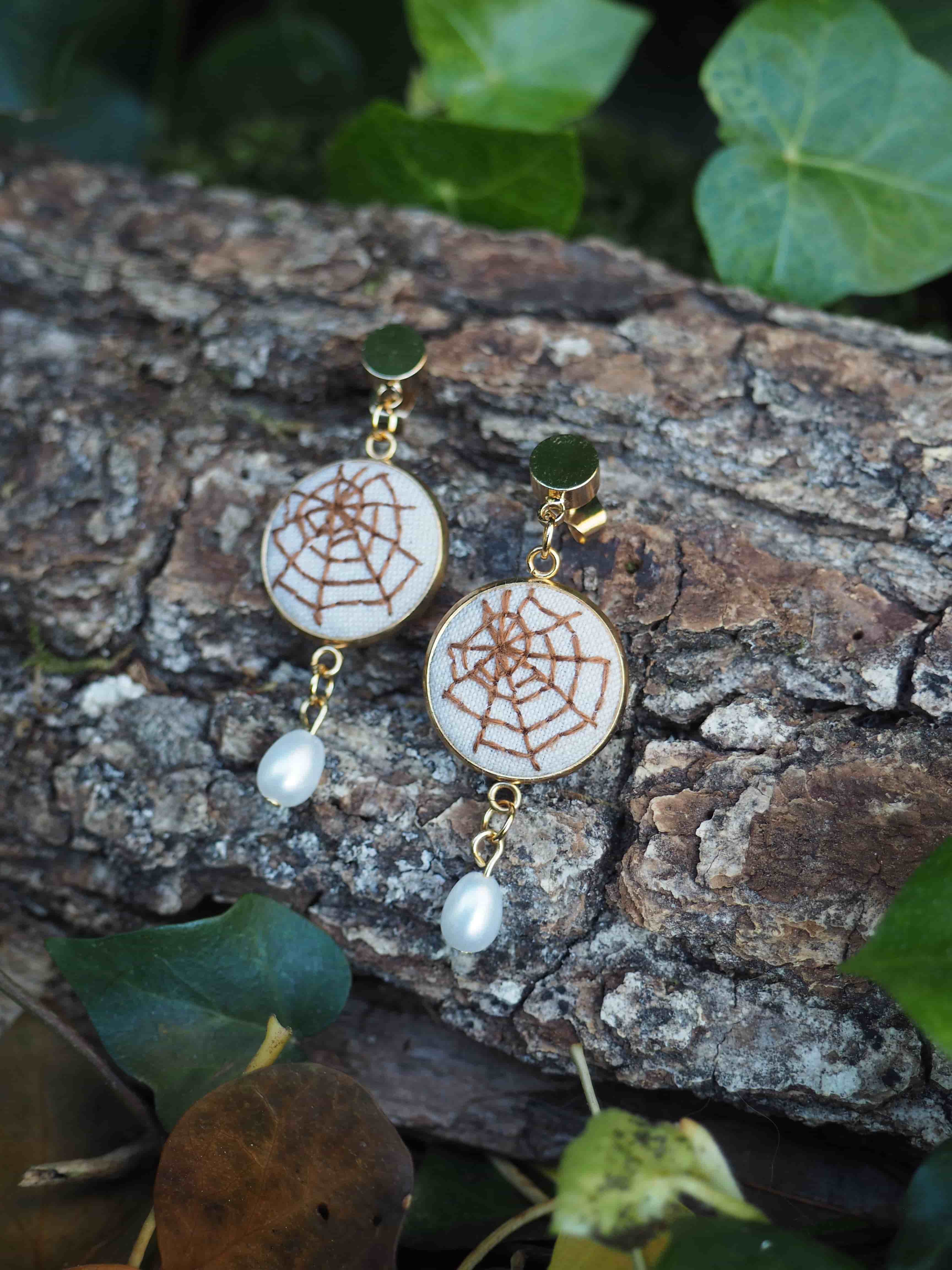 Cette image présente une paire de boucles d'oreilles élégantes disposées sur un morceau d'écorce naturelle dans un environnement végétal. **Description des bijoux :**
- Boucles d'oreilles pendantes avec des crochets dorés
- Pendentifs principaux circulaires de couleur blanche/crème avec un motif décoratif brun rappelant une toile d'araignée ou un mandala
- Petites perles blanches nacrées suspendues en bas de chaque boucle d'oreille **Mise en scène :**
- Les bijoux reposent sur un morceau d'écorce rugueuse aux tons bruns et gris
- L'arrière-plan est composé de feuillage vert, créant un contraste naturel
- L'éclairage met en valeur les détails des boucles d'oreilles et crée une ambiance douce Cette présentation met en valeur le style artisanal et naturel des bijoux, soulignant leur élégance dans un cadre organique qui complète parfaitement leur esthétique.