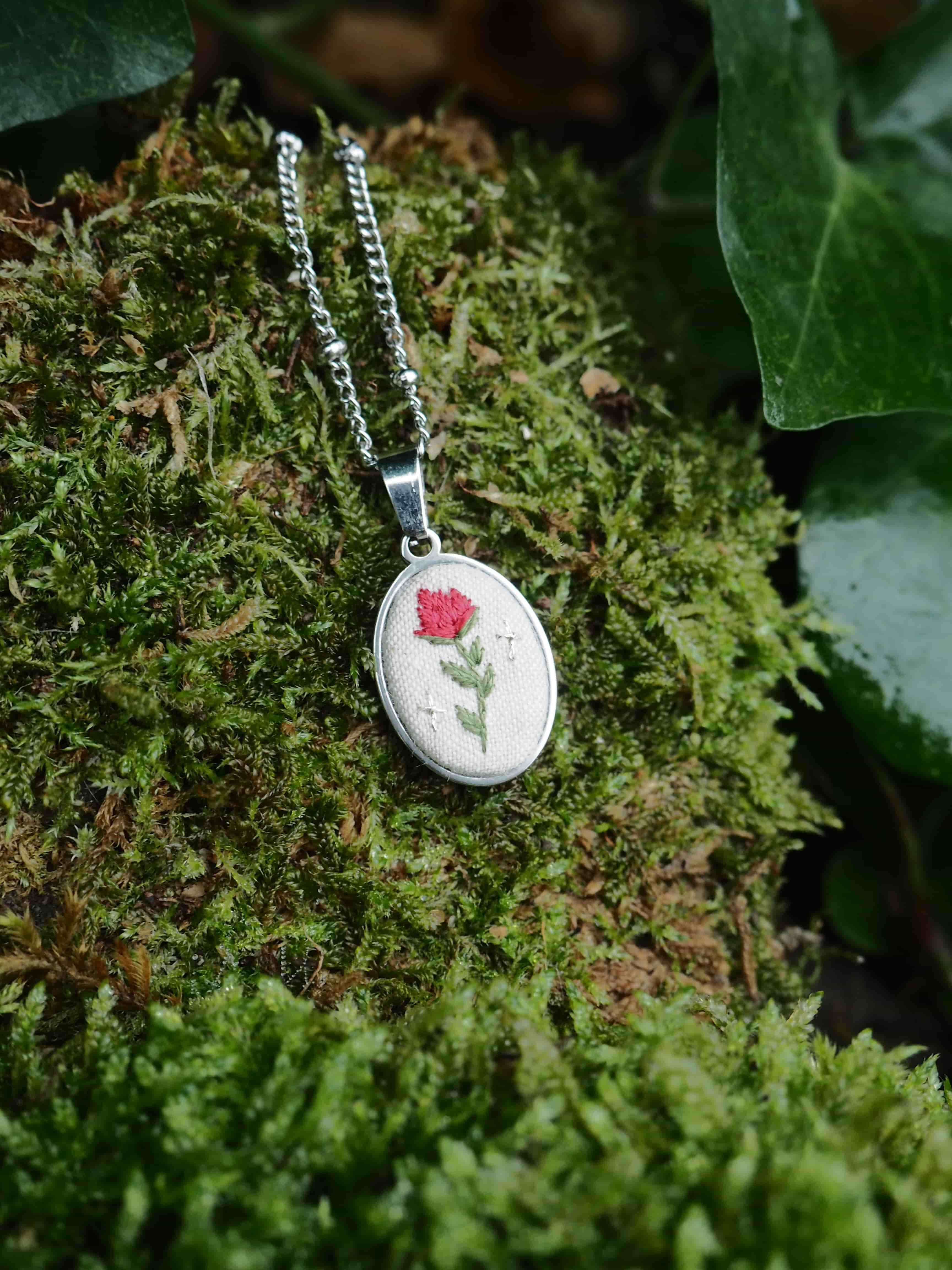 This image features a close-up shot of a delicate, oval-shaped pendant necklace resting on a bed of vibrant green moss. The pendant itself has a silver-toned metal frame, and within it, there's a light-colored, possibly cream or off-white, fabric background. Embroidered on this fabric is a charming design: a small, red flower with a green stem and leaves. To the right of the flower, there are two tiny, white embroidered star-like shapes, adding a whimsical touch. The pendant is attached to a thin, silver-toned chain, which is partially visible, extending upwards and to the left. The moss provides a natural and textured backdrop, with varying shades of green and some hints of brown underneath, suggesting depth and an organic environment. In the upper right corner and along the top edge, darker green leaves, possibly ivy, are visible, further enhancing the natural setting. The lighting appears soft, highlighting the details of the embroidery and the texture of the moss.