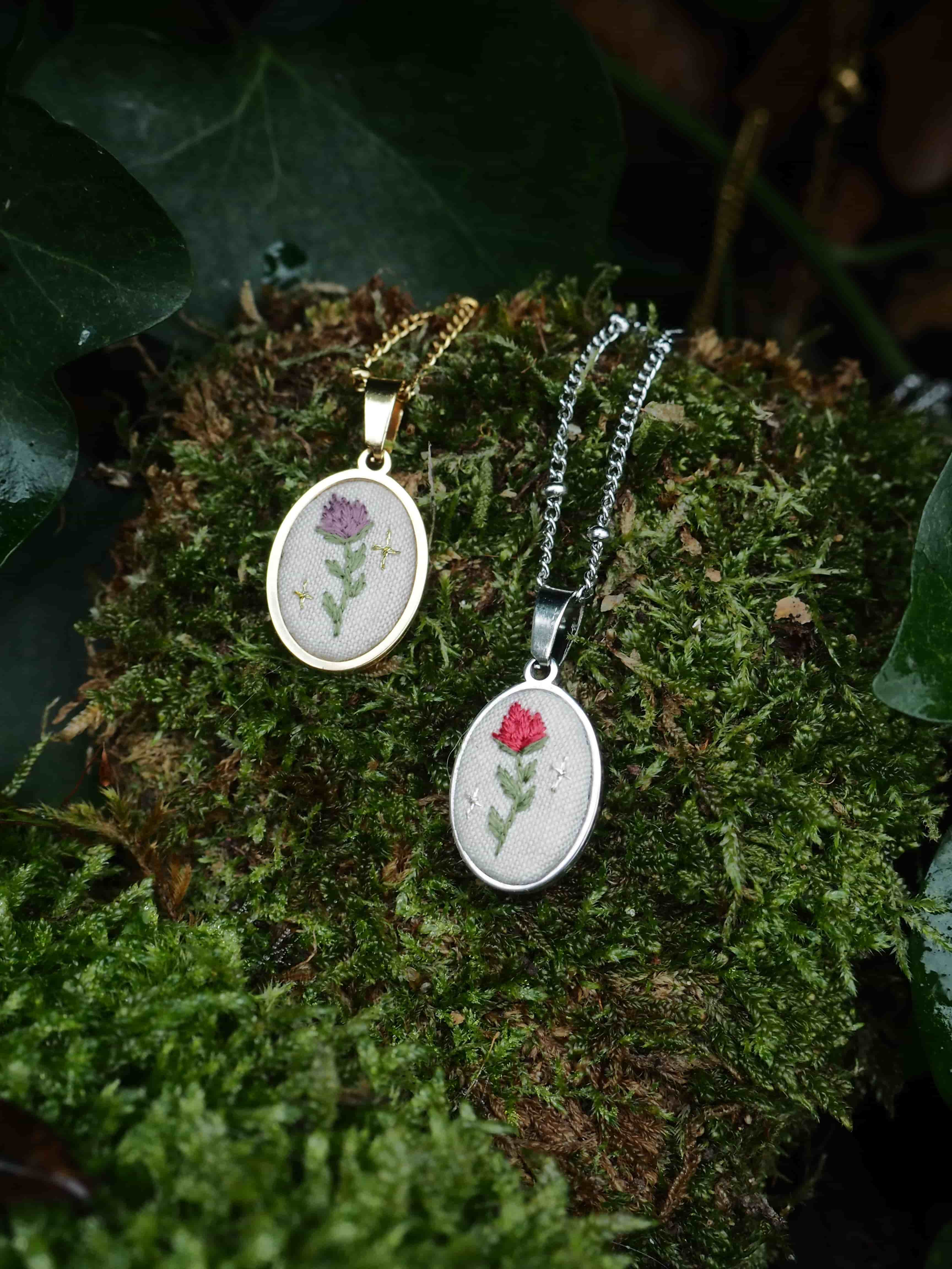 This image features two oval-shaped pendant necklaces, each showcasing a delicate embroidered floral design, set against a natural backdrop of vibrant green moss and dark green leaves. The necklace on the left has a gold-colored chain and bezel. Its pendant displays an embroidered purple flower with a green stem and leaves, accompanied by small, light-colored star-like accents on a white fabric background. The necklace on the right has a silver-colored chain and bezel. Its pendant features an embroidered red flower with a green stem and leaves, also with small, light-colored star-like accents on a white fabric background. Both necklaces are positioned on a thick bed of lush, textured green moss, which appears slightly damp in places, suggesting an outdoor or natural setting. In the background and foreground, dark green leaves, possibly ivy, are visible, some with water droplets, adding to the natural and slightly damp aesthetic of the scene. The lighting is soft, highlighting the details of the embroidery and the texture of the moss.