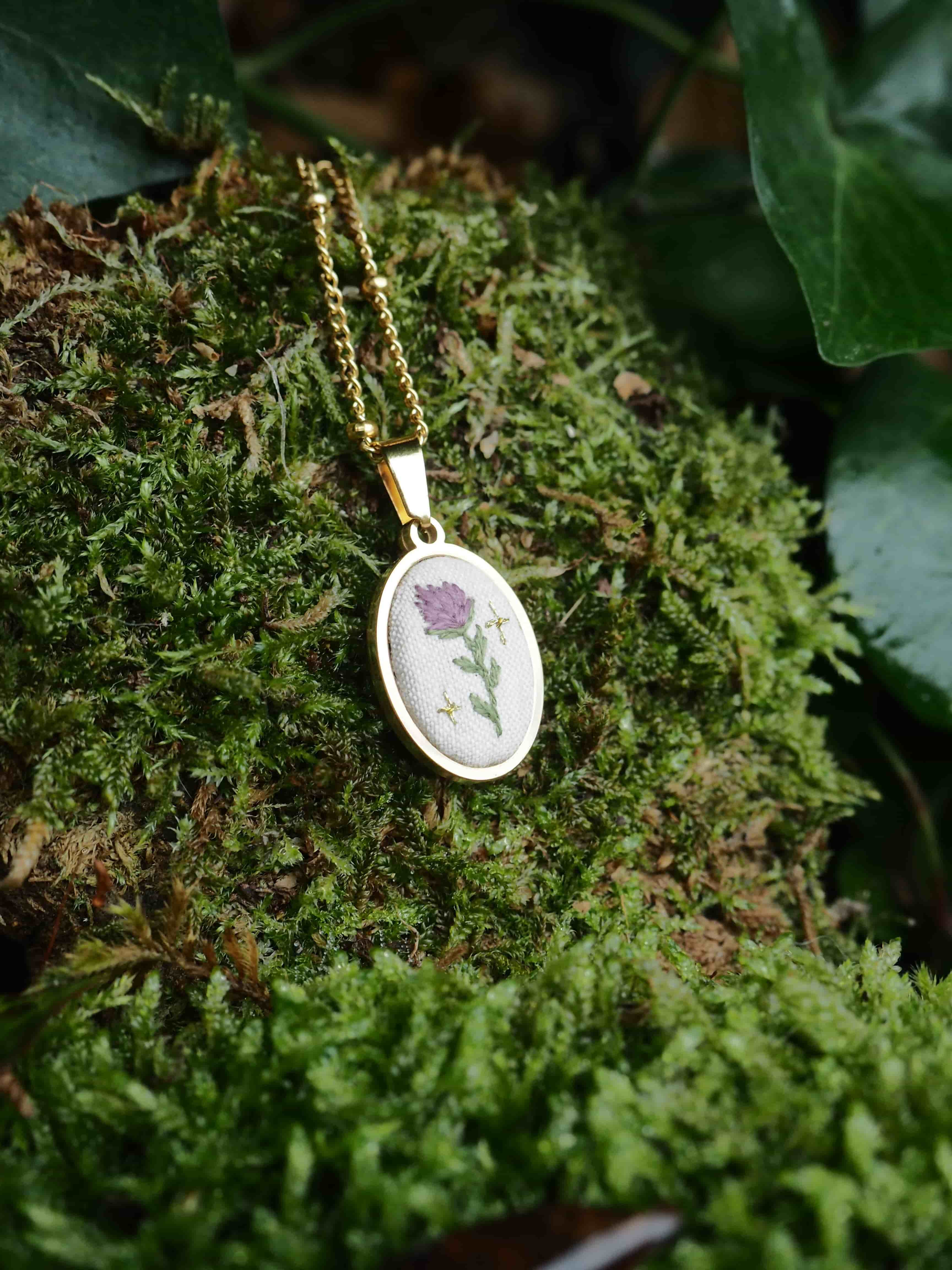 This close-up shot features a delicate gold-colored necklace with an oval pendant, resting on a bed of vibrant green moss. The pendant has a white fabric insert with a small, intricately embroidered design of a purple flower with a green stem and leaves, flanked by two tiny golden star-like accents. The gold chain of the necklace is visible, leading up from the pendant. The moss provides a soft, natural backdrop, with varying textures and shades of green, suggesting an outdoor or natural setting. In the background, hints of darker green foliage, possibly ivy, can be seen, adding depth to the image. The lighting appears soft, highlighting the details of the necklace and the texture of the moss.