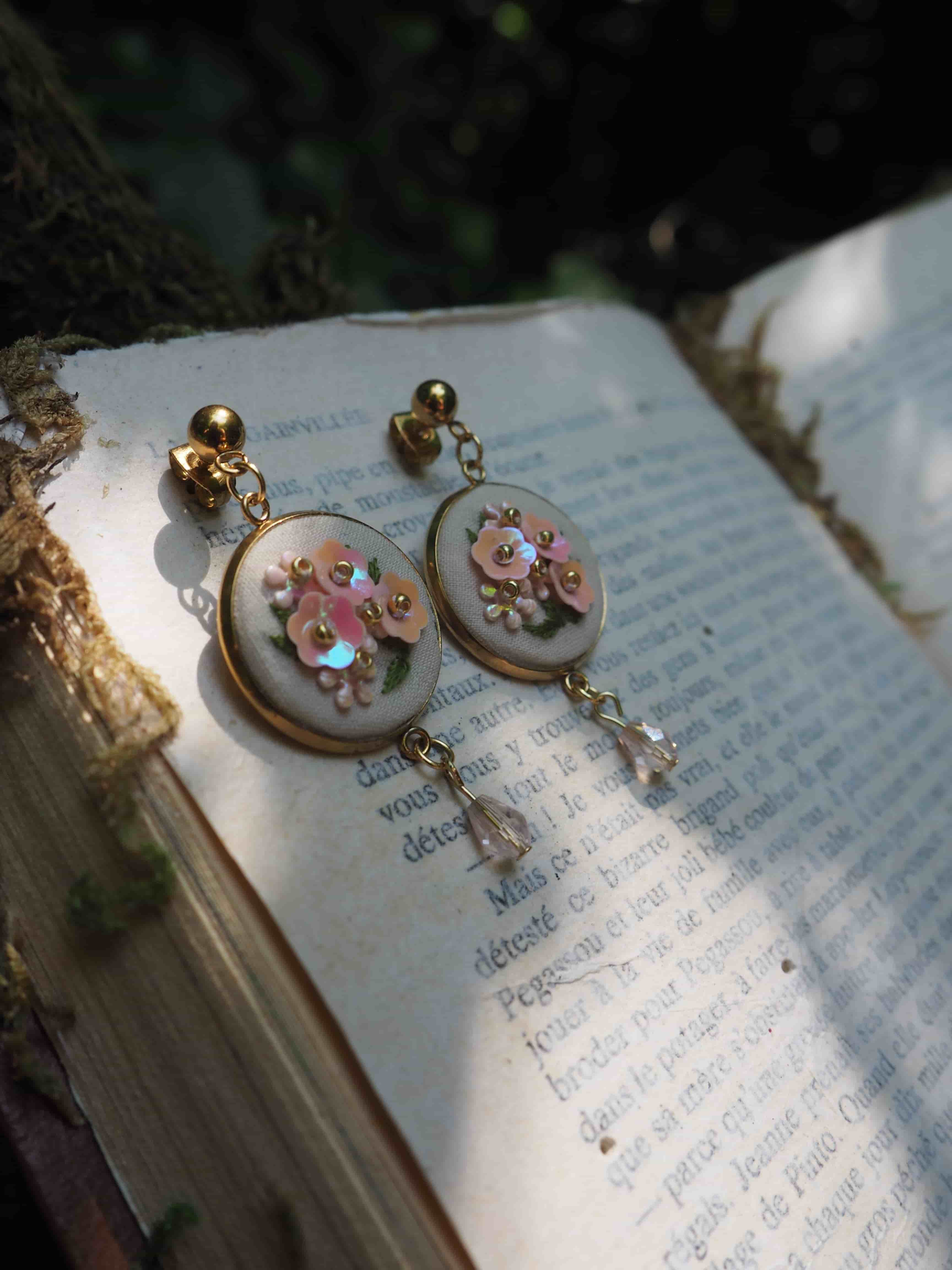 Cette image montre une paire de boucles d'oreilles délicates posées sur un livre ouvert. Les boucles d'oreilles semblent être de style vintage ou romantique, avec des médaillons ronds ornés de petites fleurs roses et de détails décoratifs. Elles sont montées sur ce qui semble être un fond nacré ou blanc, avec des petites breloques qui pendent au bas. Les boucles d'oreilles reposent sur les pages d'un livre avec du texte imprimé, créant une composition très poétique et romantique. La photo est prise avec une belle profondeur de champ qui rend l'arrière-plan légèrement flou, mettant en valeur les bijoux au premier plan.