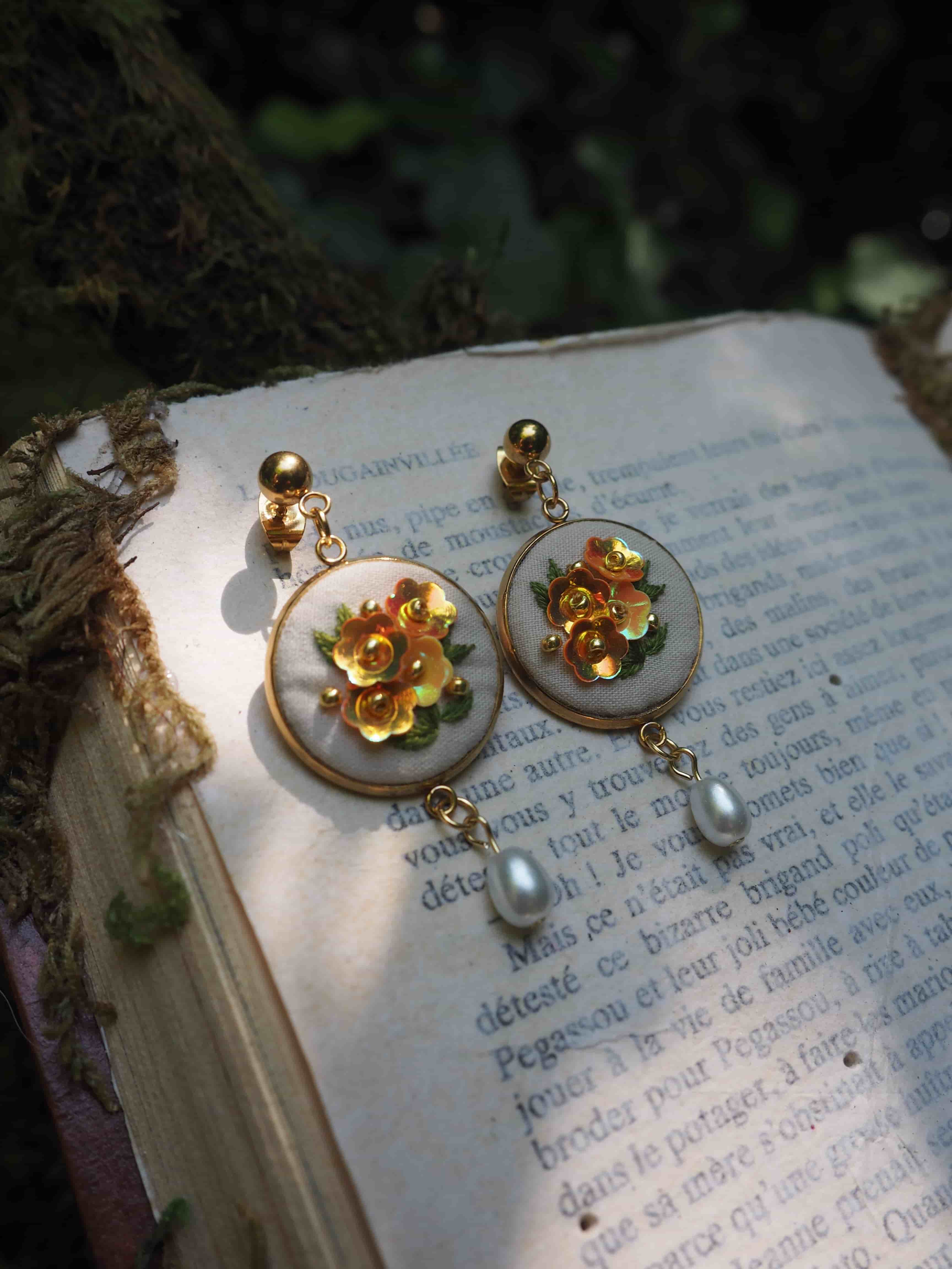 Cette image montre une paire de boucles d'oreilles élégantes posées sur les pages d'un livre en français. Les boucles d'oreilles sont de style vintage ou romantique, composées de médaillons ronds en nacre ou porcelaine blanche, ornés de fleurs dorées en leur centre. Chaque boucle d'oreille est agrémentée d'une petite perle qui pend en dessous. La photo est prise dans un éclairage naturel doux, créant une ambiance poétique et romantique. L'arrière-plan montre du texte français imprimé sur les pages du livre, ajoutant une touche littéraire et française à la composition. L'ensemble crée une atmosphère très délicate et féminine.
