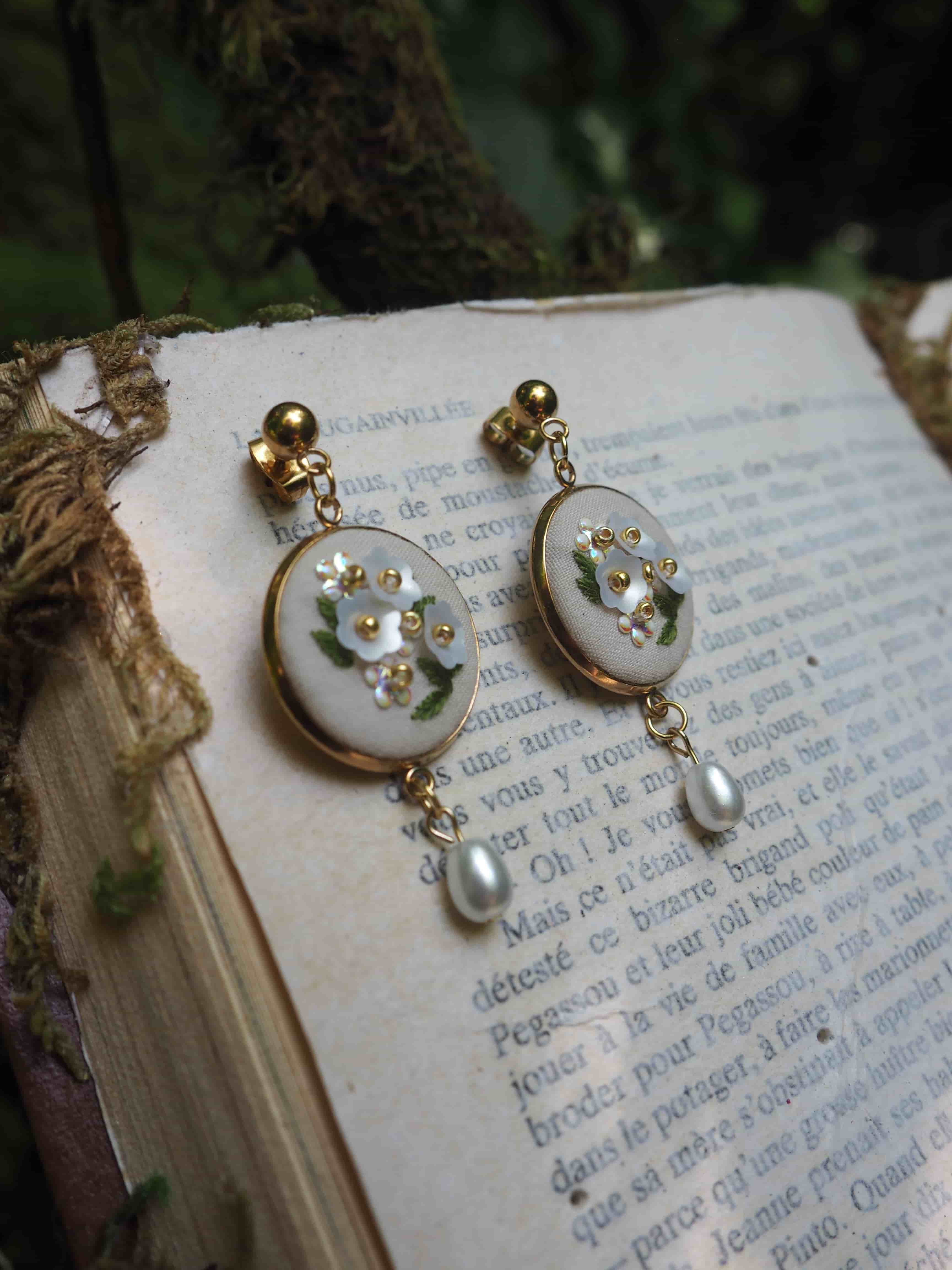 Cette image montre une paire de boucles d'oreilles élégantes posées sur les pages d'un vieux livre en français. Les boucles d'oreilles sont de style vintage avec des disques blancs ornés de petites fleurs dorées et de cristaux. Chaque boucle d'oreille se termine par une perle nacrée suspendue. Le fond montre les pages jaunies du livre avec du texte français visible, et on aperçoit ce qui semble être de l'écorce d'arbre ou du bois naturel dans le coin supérieur de l'image. La composition a un aspect très romantique et poétique, mélangeant les éléments délicats des bijoux avec la littérature classique.