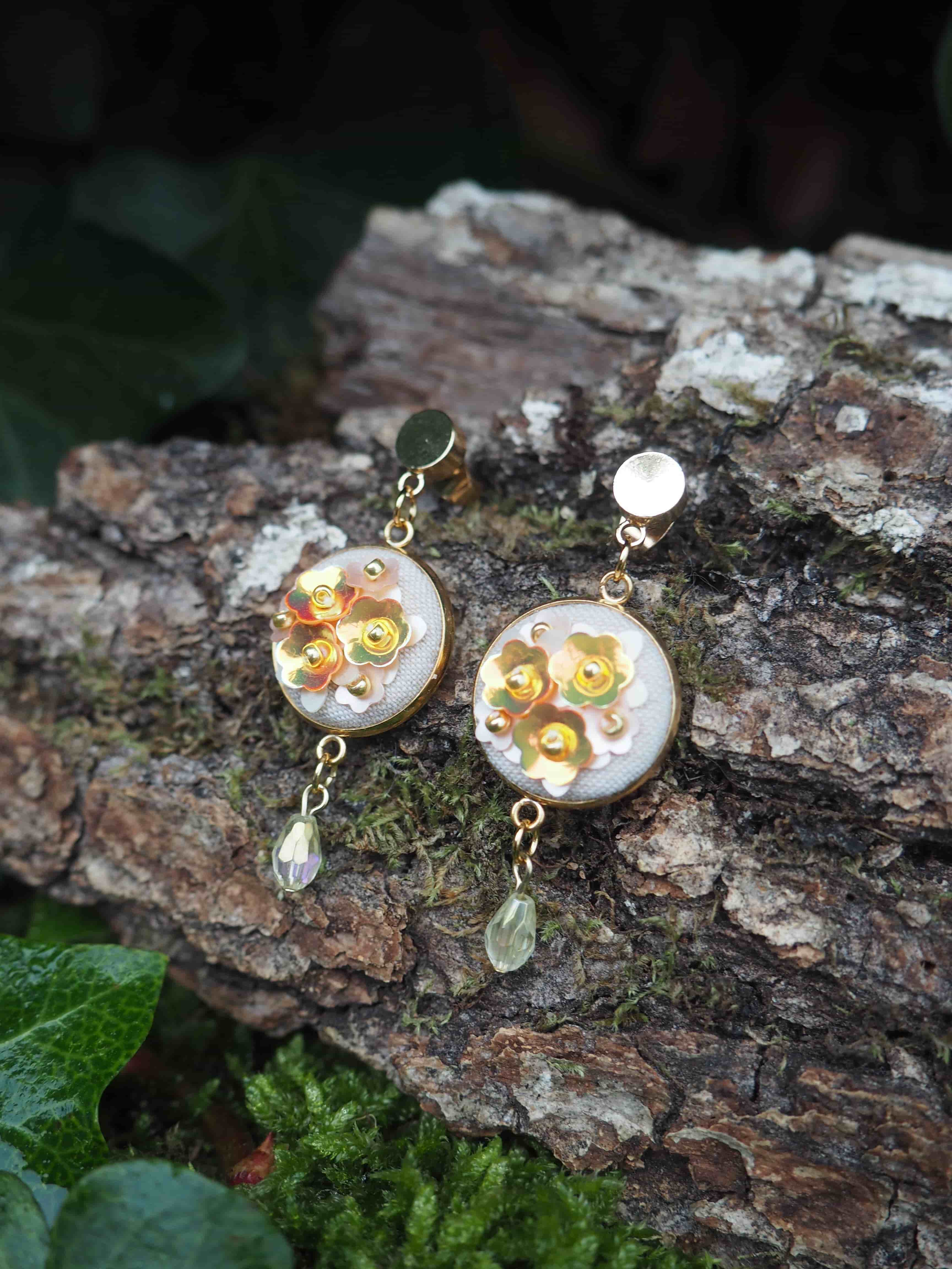 This close-up shot features a pair of elegant, handcrafted earrings resting on a textured tree bark surface, adorned with patches of green moss and hints of dark green foliage in the background. The earrings are designed with a circular, flat base, approximately 2-3 cm in diameter, framed by a thin gold-colored metal rim. The center of each circle is filled with a light, possibly off-white or very pale grey, fabric or material. Adorning this base are several small, shiny, gold-colored sequins shaped like flowers, arranged in a cluster. Some tiny, lighter-colored beads or dots are interspersed among the sequin flowers, adding to the intricate detail. Each earring dangles from a gold-colored stud or clip-on mechanism, connected by a short chain. Below the main circular element, a single, teardrop-shaped bead hangs from another small chain. The bead on the left earring appears to be a clear or iridescent light amber color, while the bead on the right earring is a translucent pale green. The lighting is soft, highlighting the metallic sheen of the gold elements and the sequins, and casting subtle shadows that emphasize the texture of the bark. The overall impression is one of delicate craftsmanship and natural beauty.
