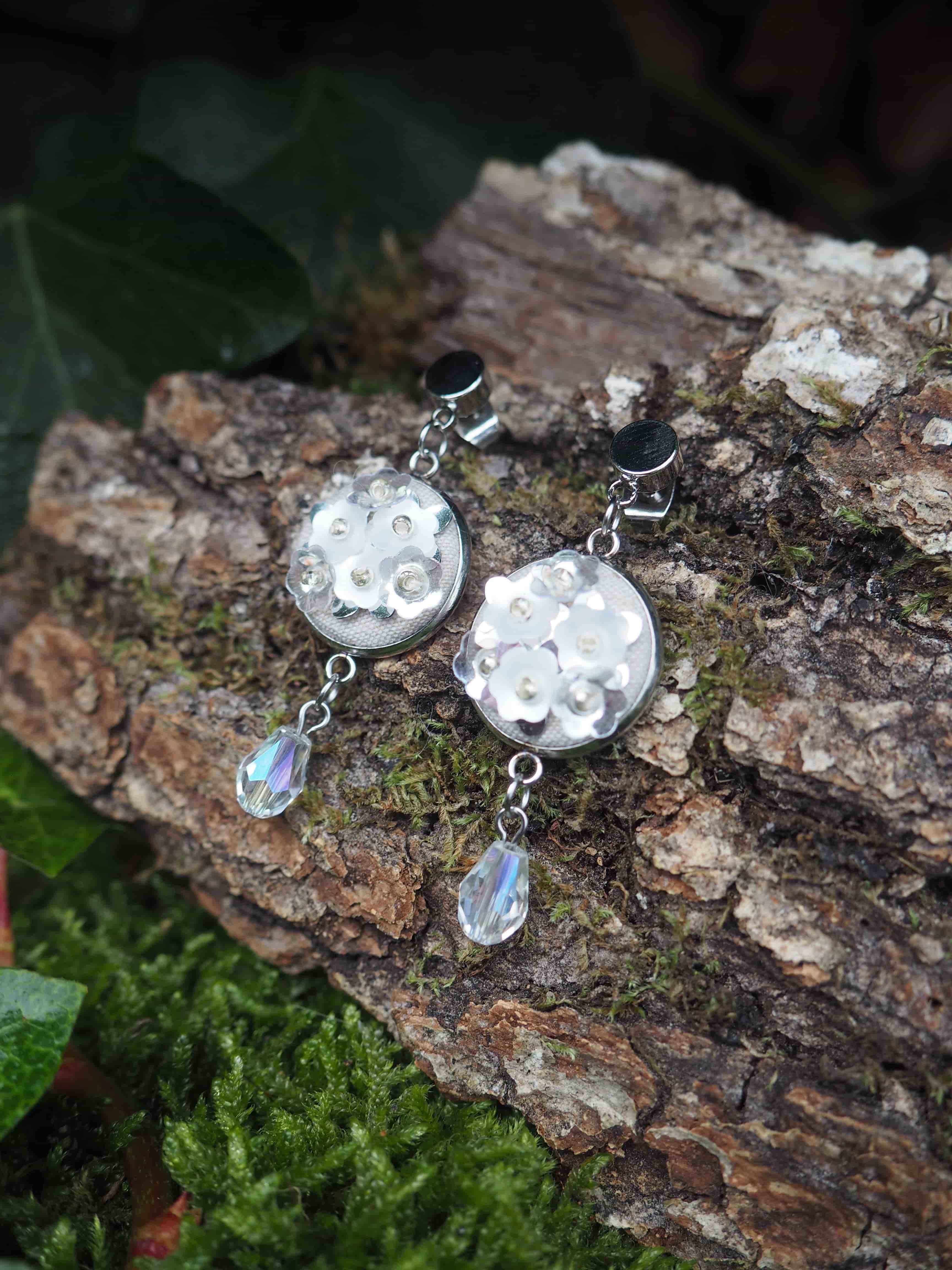 This close-up shot features a pair of elegant, dangle earrings resting on a textured piece of tree bark, which is partially covered in vibrant green moss. The natural setting provides a rustic and earthy backdrop for the delicate jewelry. Each earring consists of a circular, silver-toned base from which a clear, faceted teardrop-shaped crystal dangles. The circular part of the earrings is adorned with several small, white, opaque floral embellishments, possibly made of resin or mother-of-pearl, giving them a soft, ethereal quality. Tiny silver beads or sequins are interspersed among the flowers, adding a subtle sparkle. The earrings are attached to a silver-toned clip-on mechanism, suggesting they are designed for non-pierced ears. The tree bark is rich in texture, with deep grooves and varying shades of brown and grey, indicating its age and natural weathering. The moss growing on the bark is a bright, fresh green, contrasting beautifully with the muted tones of the wood and the shimmering white and silver of the earrings. In the background, out of focus, are hints of dark green foliage, likely ivy or similar plant life, further enhancing the natural, outdoor aesthetic of the image. The lighting appears soft and natural, highlighting the details of the earrings and the textures of the bark and moss without harsh shadows. The overall impression is one of delicate beauty set against a rugged, natural environment.