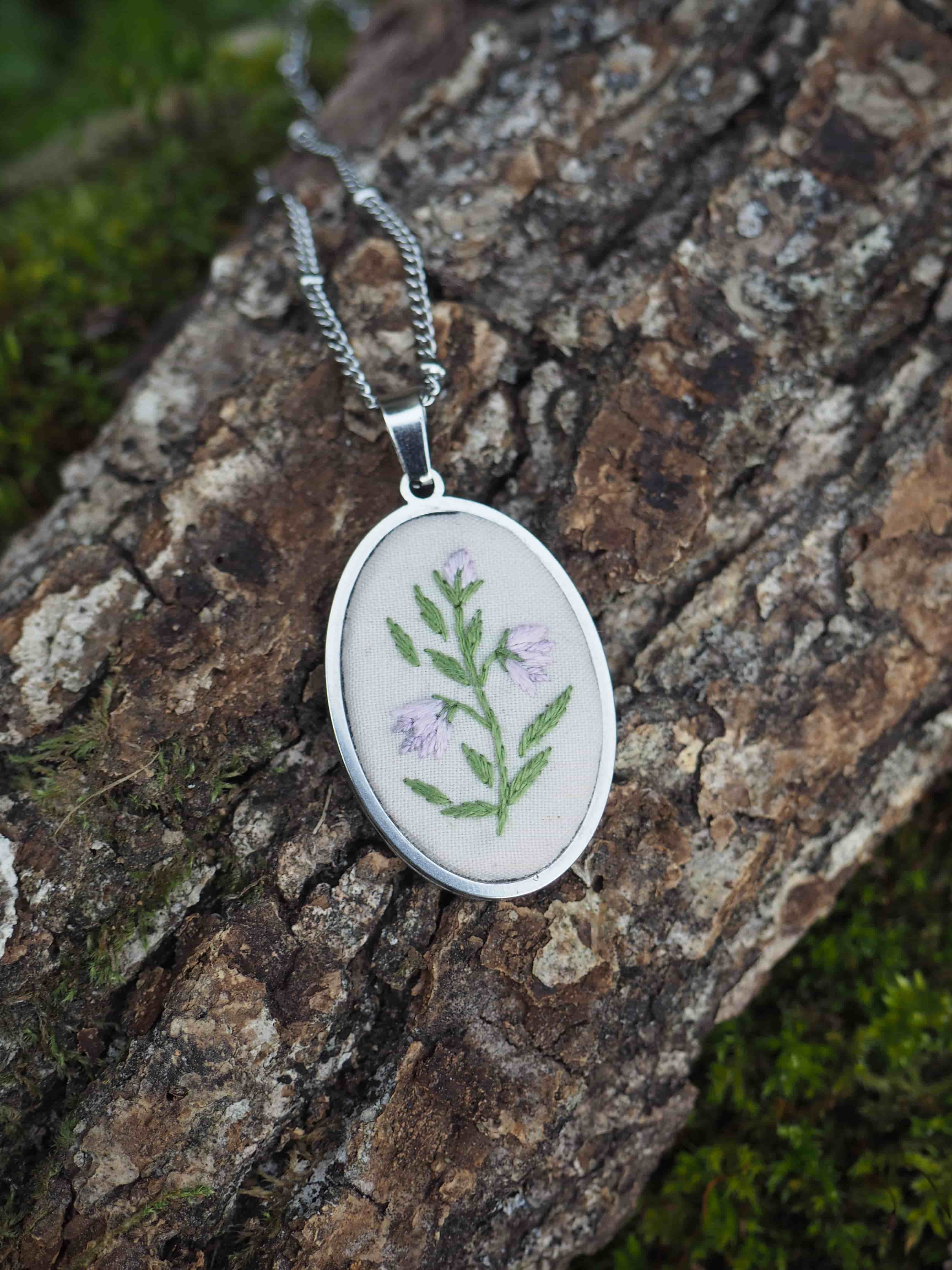 Cette image montre un pendentif délicat et artisanal. Il s'agit d'un médaillon rond en métal argenté qui contient une broderie délicate représentant une petite branche avec des feuilles vertes et de petites fleurs violettes sur un fond blanc. Le pendentif est photographié sur ce qui semble être de l'écorce d'arbre texturée avec un peu de mousse verte, créant un beau contraste naturel avec le bijou. Le pendentif est attaché à une fine chaîne argentée. C'est un très beau exemple de bijou qui combine l'artisanat textile traditionnel (la broderie) avec la joaillerie.