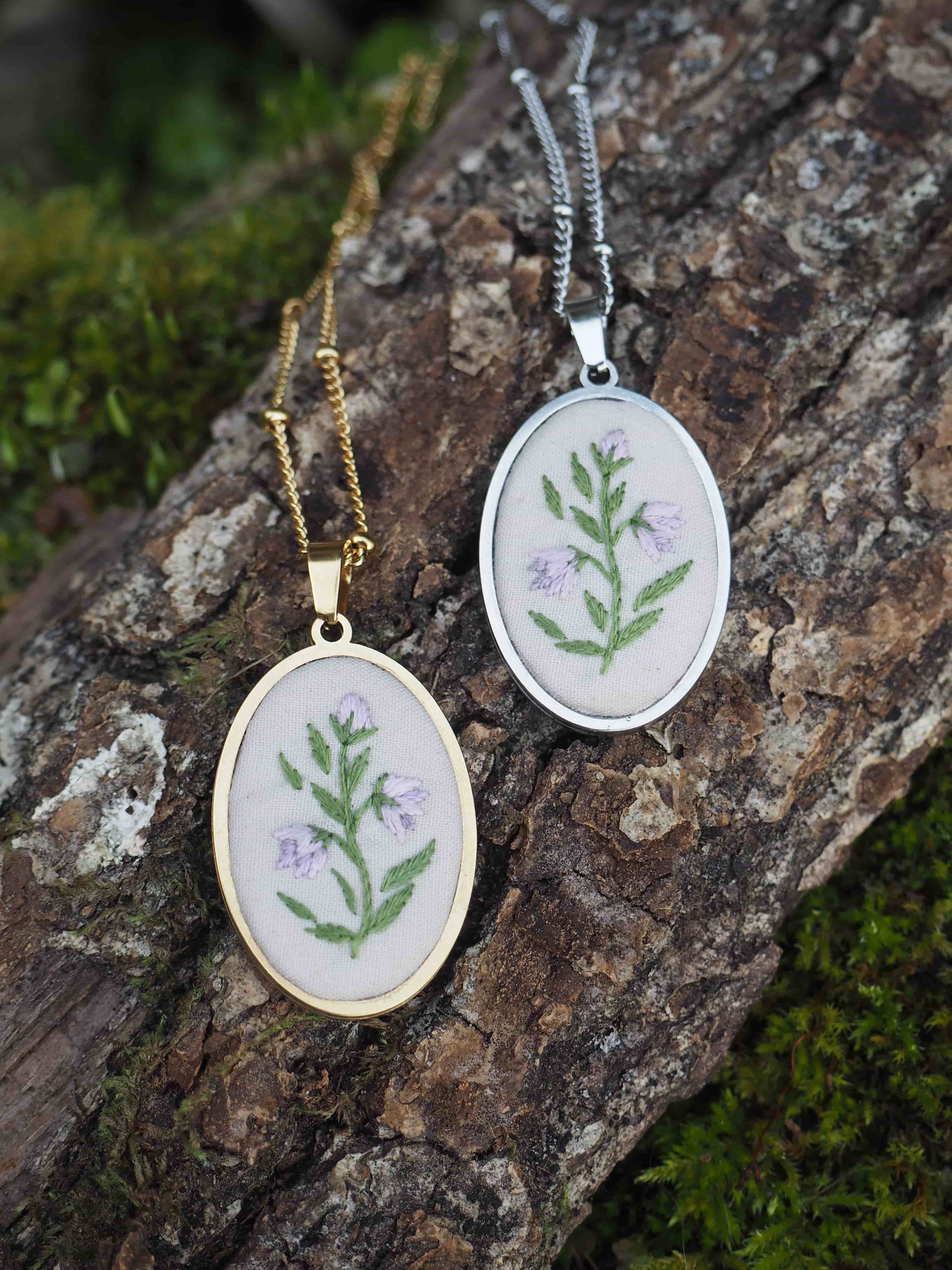 Cette image montre deux pendentifs délicats posés sur un morceau de bois ou d'écorce recouvert de mousse. Les pendentifs sont des médaillons circulaires, l'un en métal doré et l'autre en argent, chacun contenant une broderie botanique. La broderie représente une petite branche avec des feuilles vertes et ce qui semble être de petites fleurs violettes, réalisée sur un fond de tissu clair. Les pendentifs sont attachés à des chaînes fines assorties à leur couleur respective. La composition est très naturelle et met en valeur ces bijoux artisanaux dans un cadre organique.
