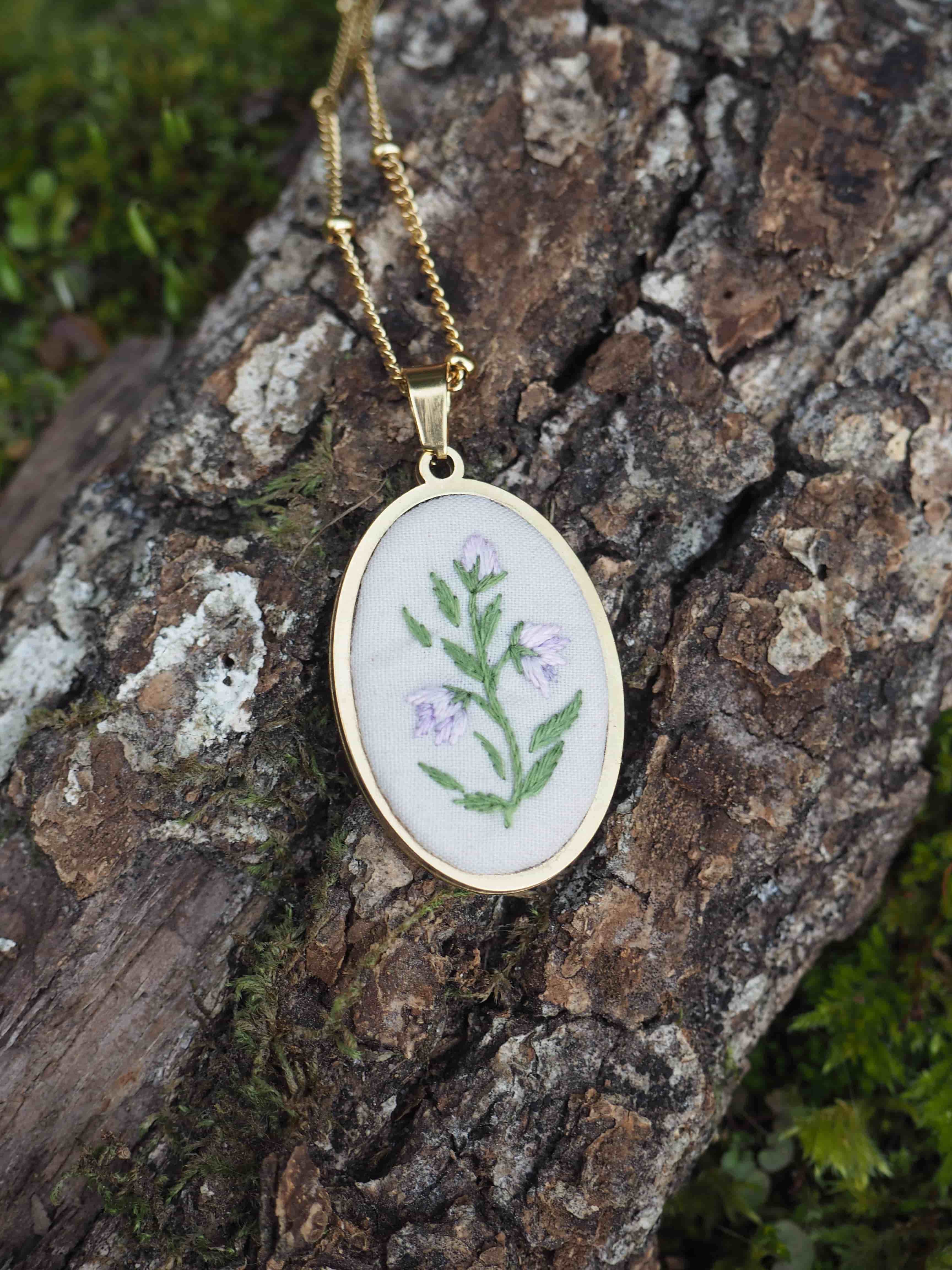 Cette image montre un délicat pendentif de broderie placé sur un morceau d'écorce d'arbre texturée et moussue. Le pendentif est rond, serti dans un cadre doré, et présente une broderie sur fond blanc avec un motif floral simple - des feuilles vertes et de petites fleurs violettes. Il est suspendu à une fine chaîne dorée. Le contraste entre la délicatesse du bijou artisanal et la texture rugueuse de l'écorce naturelle crée une belle composition photographique.