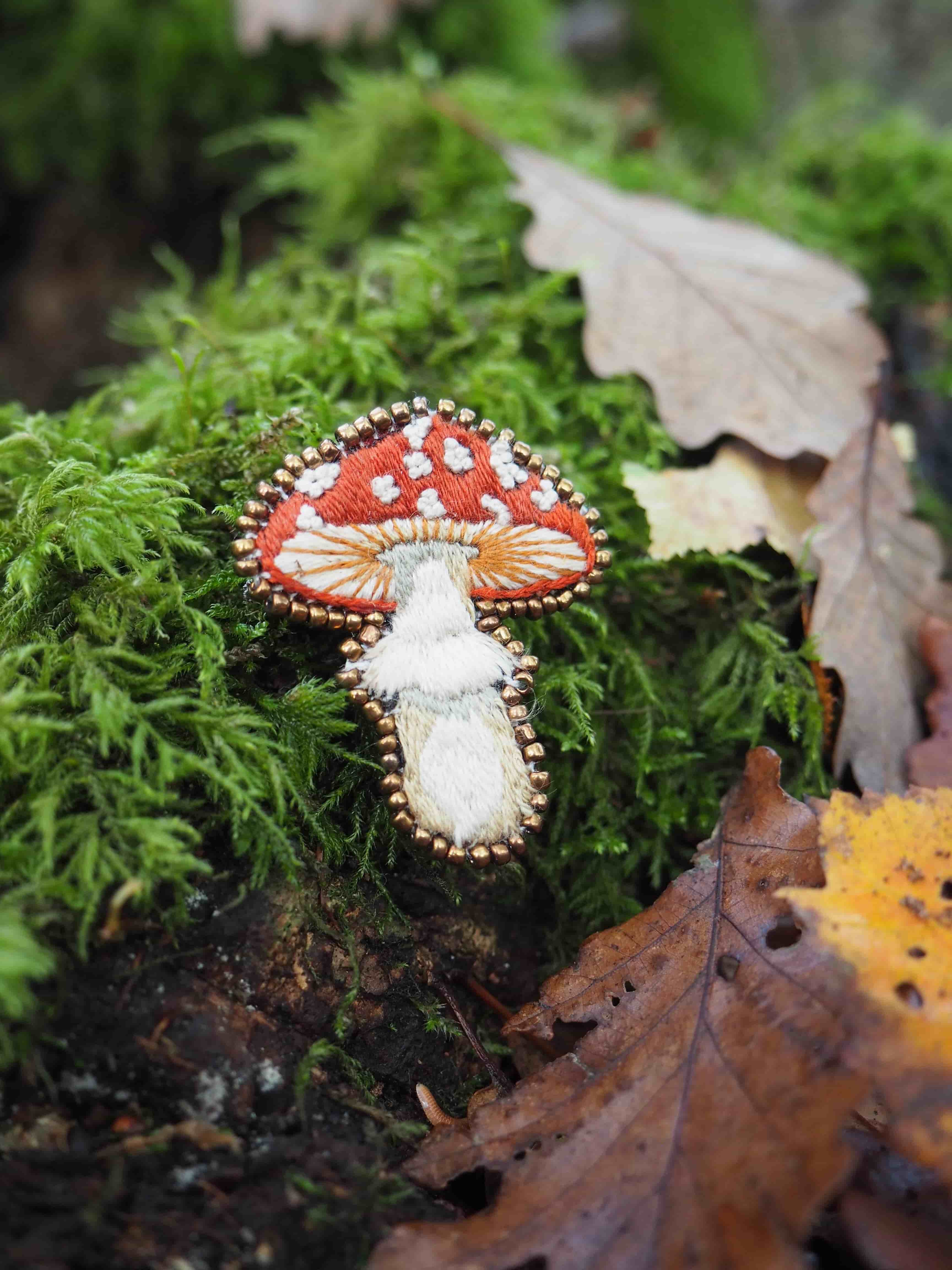 Cette image présente une scène charmante de la nature avec un élément décoratif artistique. Au premier plan, on peut voir un champignon décoratif minutieusement brodé ou cousu, représentant un champignon rouge à pois blancs (rappelant l'amanite tue-mouches) avec un pied blanc. Cette pièce artisanale est délicatement posée sur un lit de mousse verte luxuriante. L'environnement naturel qui l'entoure est composé de :
- Mousse épaisse et verdoyante qui tapisse le sol
- Feuilles mortes aux teintes automnales (brunes et dorées) éparpillées
- Une végétation forestière dense et verte en arrière-plan
- L'ensemble créant une atmosphère de sous-bois humide et paisible L'effet artistique est renforcé par une profondeur de champ qui maintient le champignon brodé net au premier plan tandis que l'arrière-plan reste légèrement flou, créant un contraste saisissant entre l'art textile et la nature authentique. Cette composition évoque un conte de fées ou un monde fantastique où l'artisanat se mêle harmonieusement à l'environnement forestier.