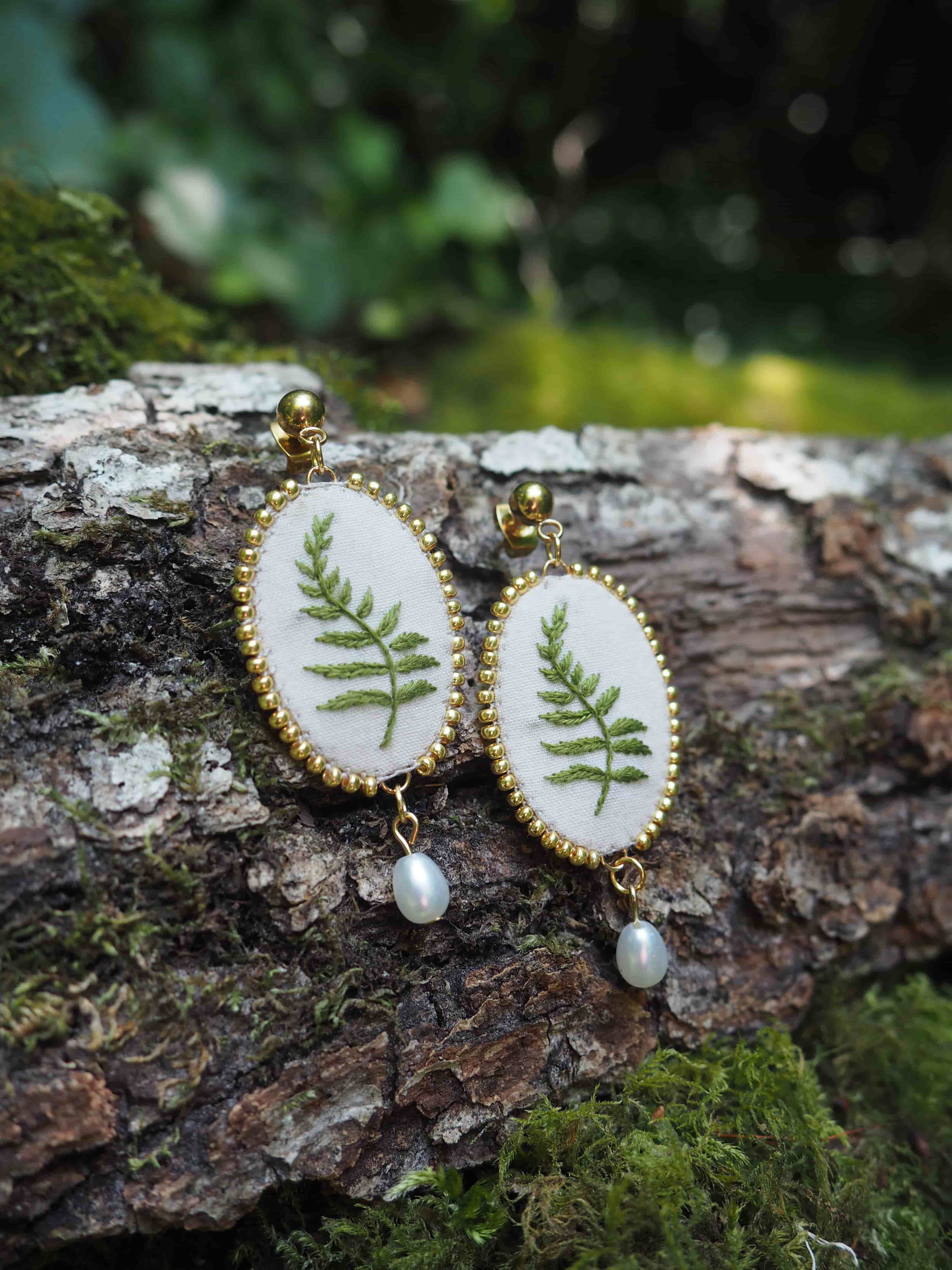 Cette image montre une paire de boucles d'oreilles artisanales très délicates. Chaque boucle d'oreille est composée d'un médaillon ovale avec un cadre doré décoratif. À l'intérieur de chaque médaillon, on peut voir une véritable feuille de fougère pressée sur un fond blanc. Au bas de chaque boucle d'oreille pend une petite perle blanche. Les boucles d'oreilles sont photographiées sur ce qui semble être un morceau d'écorce d'arbre couvert de mousse, créant un cadre naturel qui complimente parfaitement le motif botanique des bijoux. L'arrière-plan est flou mais montre des tons verts naturels, suggérant un environnement forestier. Ce sont des bijoux qui capturent vraiment l'essence de la nature, combinant élégamment des éléments naturels avec un design raffiné.