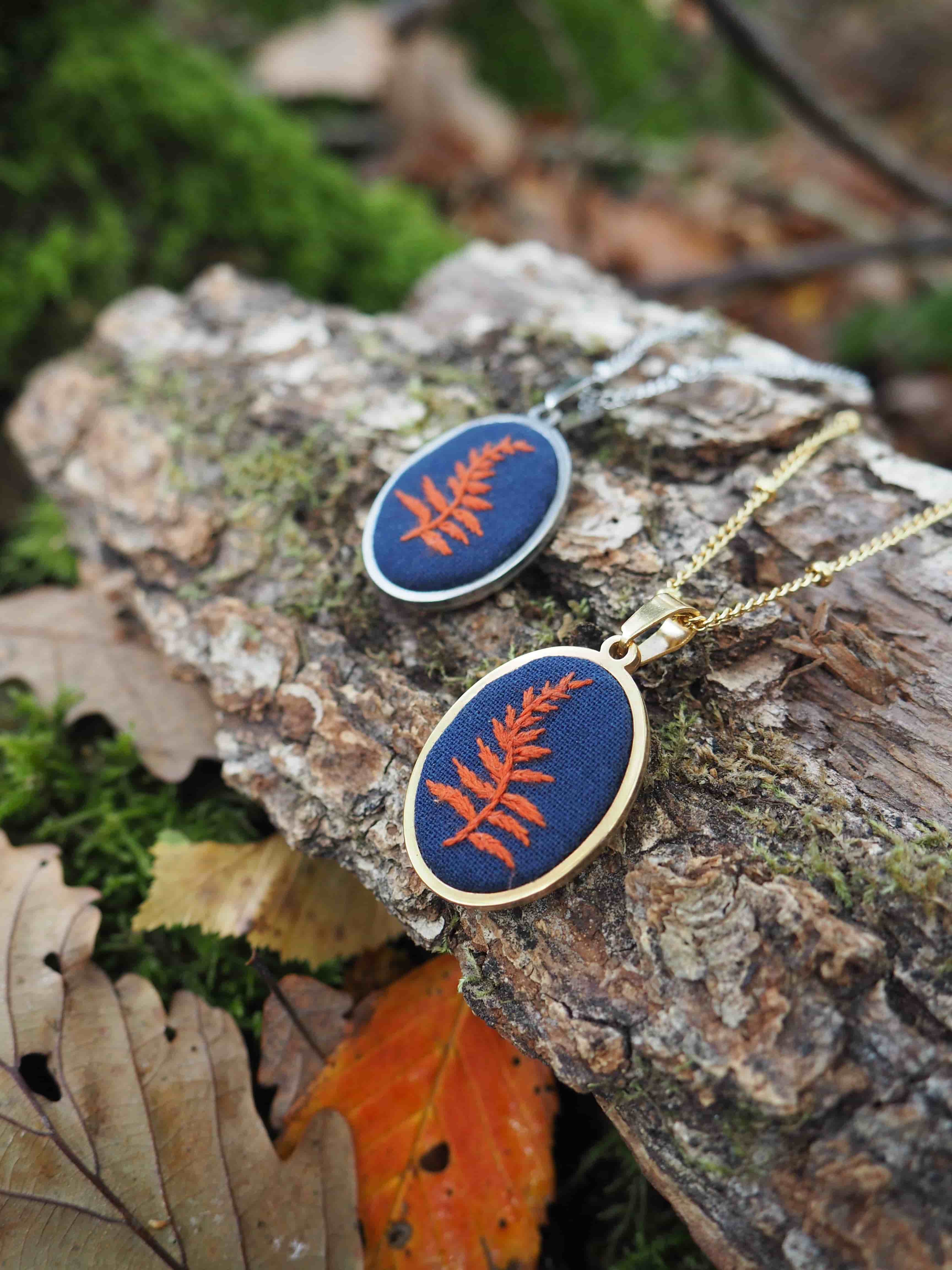 Cette image présente deux bijoux élégants posés sur un tronc d'arbre dans un environnement naturel. Il s'agit de pendentifs ou médaillons de forme ovale, montés sur des chaînes dorées délicates. **Caractéristiques des bijoux :**
- Forme ovale avec un cadre doré
- Fond bleu marine profond
- Motif central : une fougère ou feuille stylisée de couleur cuivre/orange
- Finition qui semble être de la résine ou de l'émail
- Chaînes fines de couleur dorée **Mise en scène :**
- Posés sur l'écorce rugueuse d'un tronc d'arbre
- Environnement forestier avec de la mousse verte visible
- Feuilles d'automne aux tons orangés parsemées autour
- Éclairage naturel qui met en valeur les couleurs et textures L'ensemble crée une harmonie parfaite entre les bijoux et leur environnement naturel, soulignant le thème botanique du motif de fougère. La photographie met bien en valeur le contraste entre les matériaux précieux des bijoux et la texture brute de l'écorce.