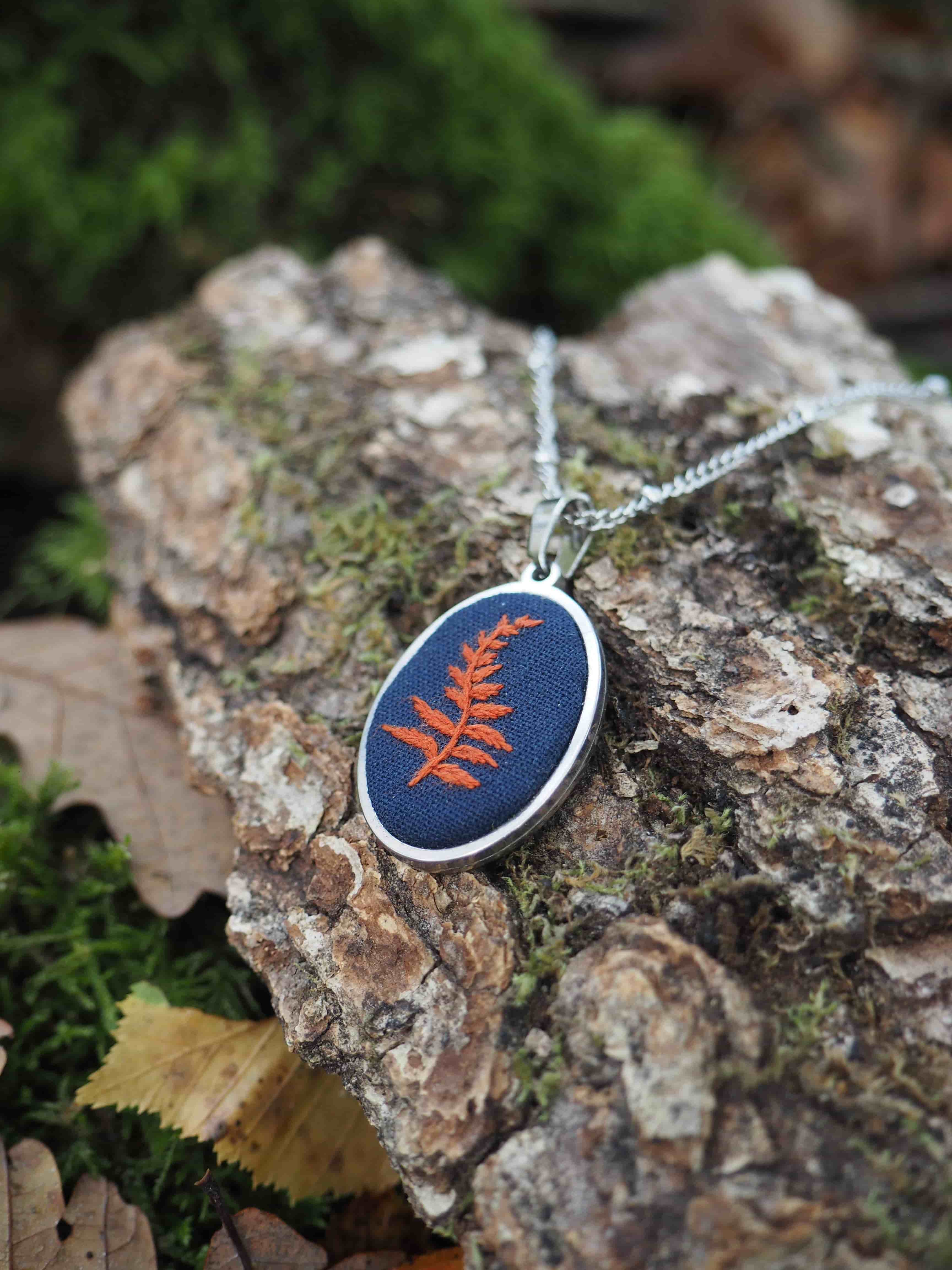 Cette image présente un pendentif artisanal posé sur un tronc d'arbre moussu dans un environnement naturel. Le bijou se compose d'un médaillon ovale avec un cadre en métal argenté, contenant un tissu ou une broderie de couleur bleu marine sur lequel est représentée une fougère ou une plante stylisée de couleur orange/rouge-orangé. Le pendentif est suspendu à une chaîne fine de couleur argentée. Il repose sur l'écorce rugueuse d'un tronc d'arbre partiellement recouvert de mousse verte, créant un contraste naturel avec l'objet artisanal. L'arrière-plan est artistiquement flouté, laissant entrevoir de la végétation verte luxuriante, ce qui donne une atmosphère forestière et naturelle à la composition. L'éclairage naturel met en valeur les détails du bijou tout en créant une ambiance douce et organique. Cette mise en scène suggère un style de bijouterie inspiré de la nature, probablement fait main, où l'artisan a voulu capturer l'essence botanique dans une pièce portable.