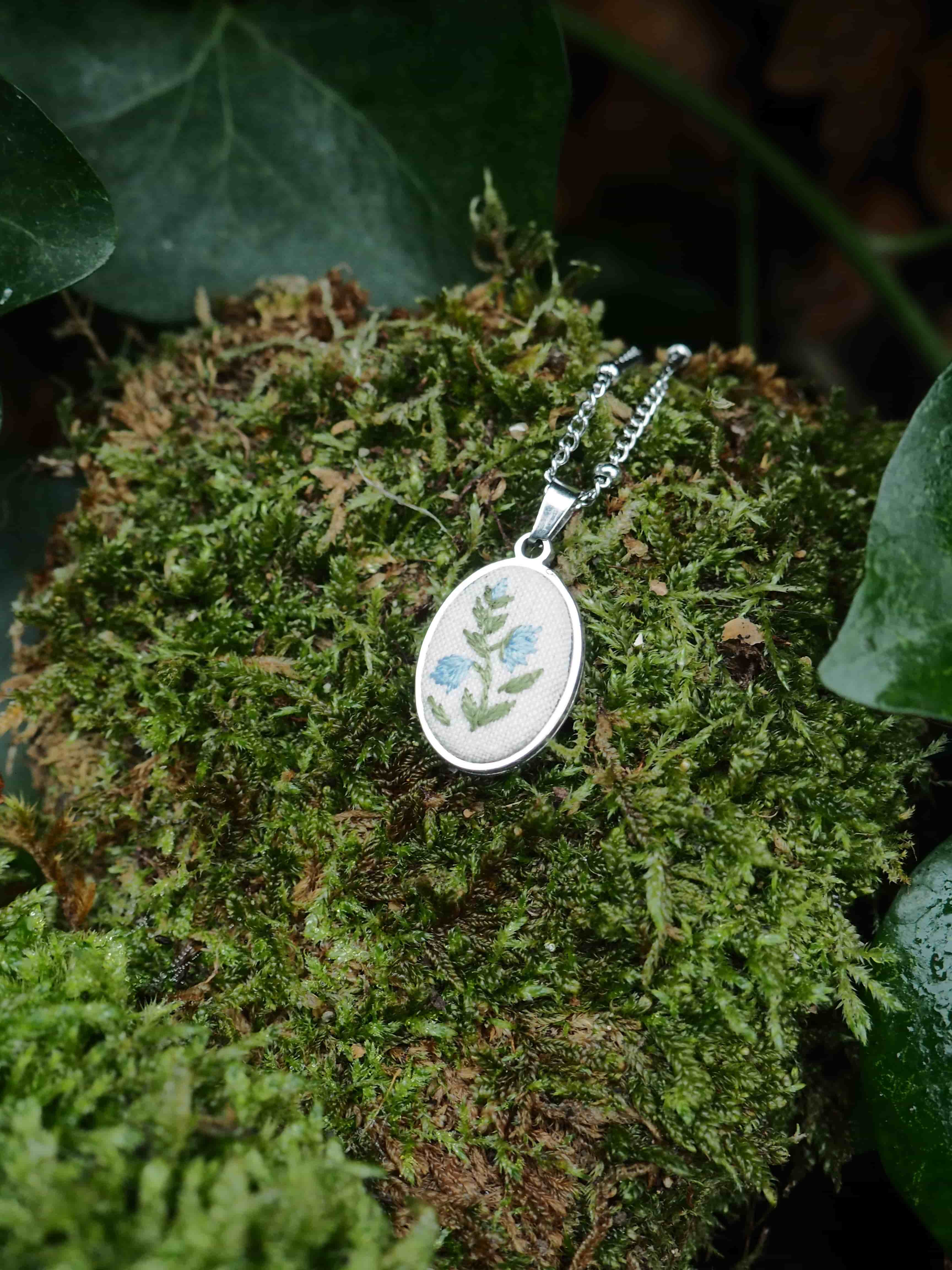 This image features a close-up shot of an oval-shaped pendant with a silver-colored chain, resting on a bed of vibrant green moss. The pendant itself has a white or light-colored background with an embroidered or painted design of light blue flowers with green stems and leaves. The design appears delicate and natural. The moss provides a textured, organic backdrop, with varying shades of green and some darker, possibly brown, undertones suggesting depth and natural growth. In the upper left and lower right corners, out-of-focus dark green leaves, possibly ivy, frame the scene, adding to the natural and earthy aesthetic. The lighting highlights the pendant and the moss, creating a soft, natural feel.