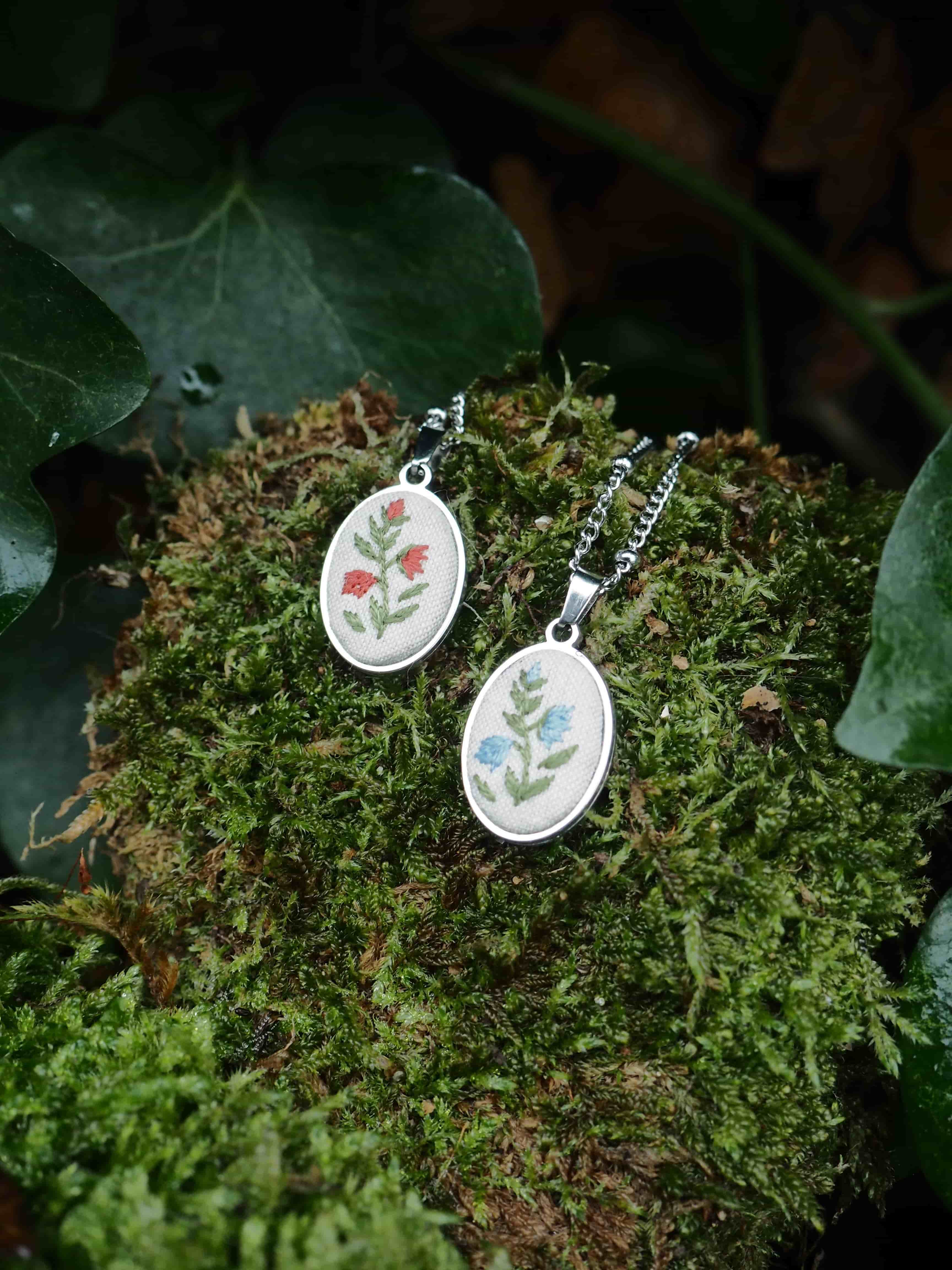 This image features two oval-shaped pendants, each with a delicate embroidered floral design, resting on a bed of vibrant green moss. The pendants are silver-toned and have a white fabric base for the embroidery. The pendant on the left displays a design of two red flowers with green stems and leaves. The pendant on the right showcases a design of two blue flowers with green stems and leaves. Both pendants are attached to silver-colored chains, which are partially visible. The moss provides a natural and textured backdrop, with varying shades of green and some brown undertones, suggesting a forest or garden setting. In the background, out of focus, are dark green leaves, possibly ivy, further enhancing the natural aesthetic of the scene. The lighting appears soft, highlighting the details of the pendants and the texture of the moss.