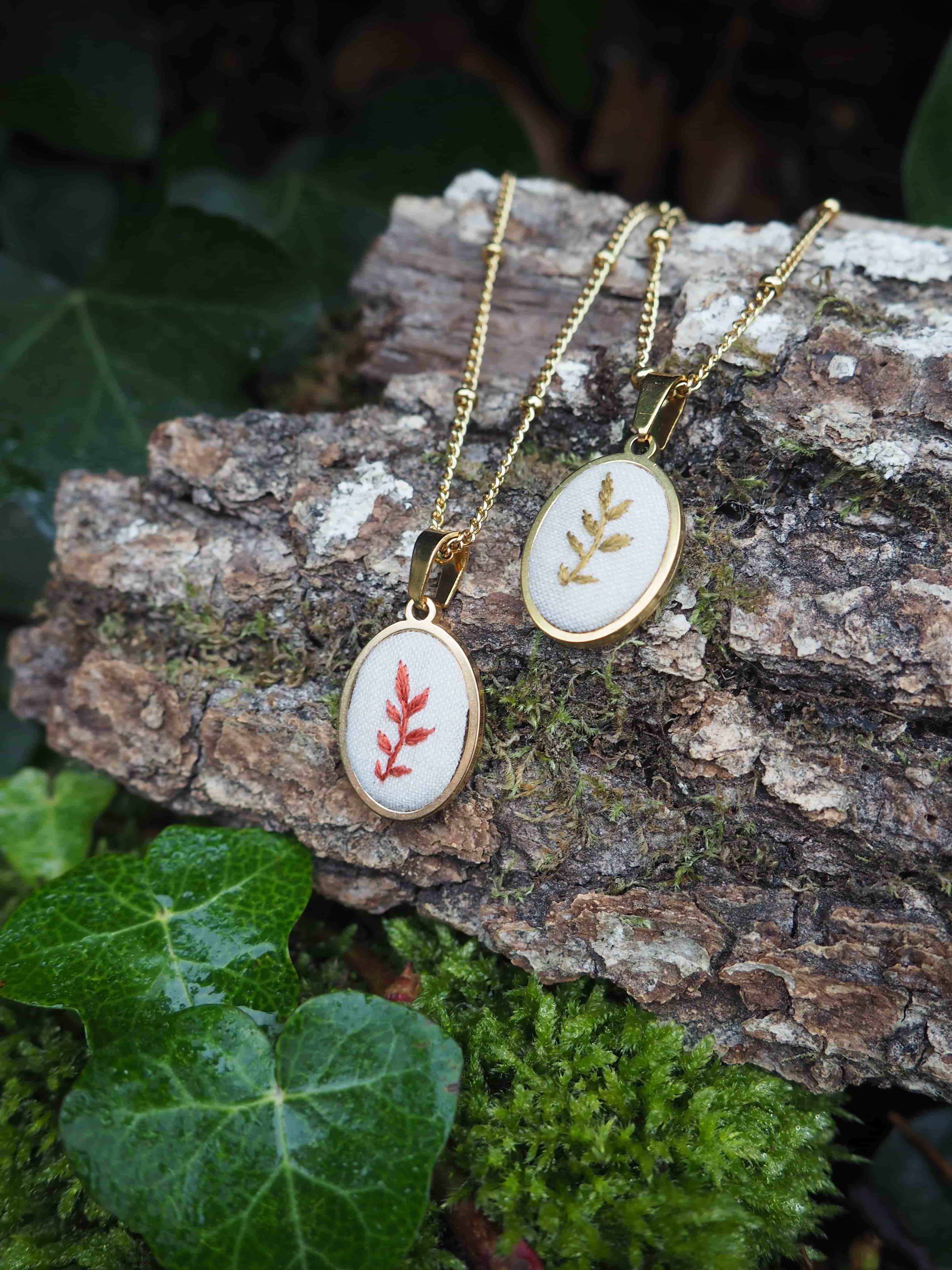 This image features two delicate oval-shaped pendants with gold-colored chains, resting on a piece of textured tree bark. The pendants are identical in shape and size, each featuring a white fabric insert with an embroidered leaf design. The pendant on the left displays a reddish-brown leaf design, while the one on the right has a golden-yellow leaf design. Both pendants are framed by a shiny gold-colored bezel. The chains are thin and have small, evenly spaced beads along their length. The tree bark beneath the pendants is rough and dark brown, with visible grooves and some patches of green moss. To the left and bottom of the pendants, there are vibrant green ivy leaves, some of which appear to have water droplets on them, suggesting a damp or natural outdoor setting. The background is softly blurred, showing hints of darker foliage. The overall impression is one of natural beauty and handcrafted elegance, with the jewelry harmonizing with its organic surroundings.