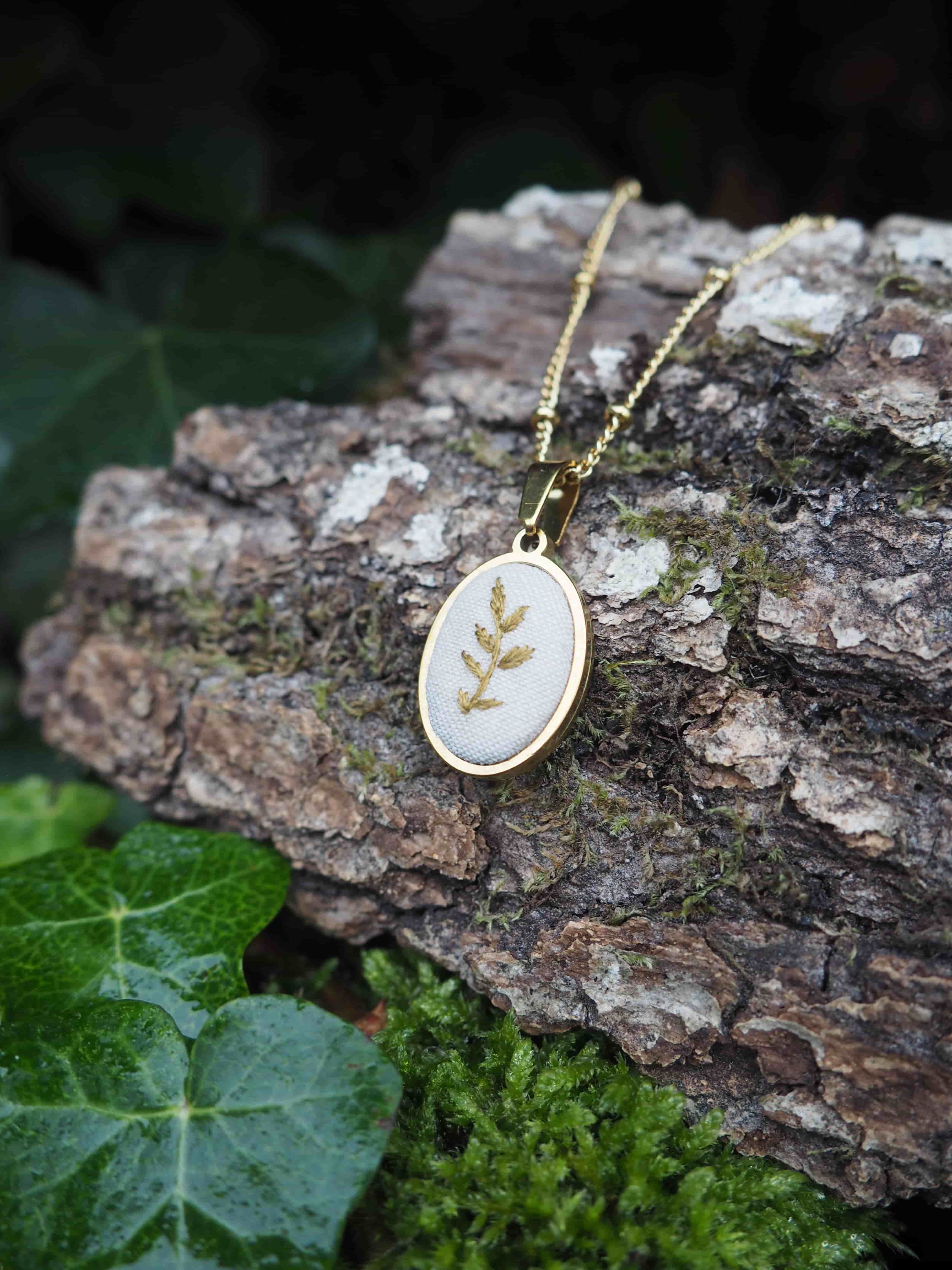 This close-up shot features a delicate gold-colored necklace with an oval pendant, resting on a piece of textured tree bark. The pendant is the focal point, showcasing a light, possibly white or cream-colored, fabric or enamel base. Embroidered or depicted on this base is a stylized golden leaf or sprig design, adding a touch of nature-inspired elegance. The pendant is framed by a thin gold-colored bezel. The necklace chain is also gold-colored and appears to be a fine, beaded or delicate link chain, partially visible as it drapes across the bark. The tree bark provides a rustic and natural backdrop, with its rough texture and varying shades of brown and grey. Patches of vibrant green moss are visible on the bark, enhancing the organic feel of the scene. In the foreground and to the left, lush green ivy leaves are visible, some with a slight sheen, suggesting they might be damp or recently watered. The background is softly blurred, with hints of dark green foliage, further emphasizing the natural setting. The lighting appears soft and natural, highlighting the metallic gleam of the necklace and the intricate details of the pendant, while creating a gentle contrast with the earthy tones of the bark and foliage.
