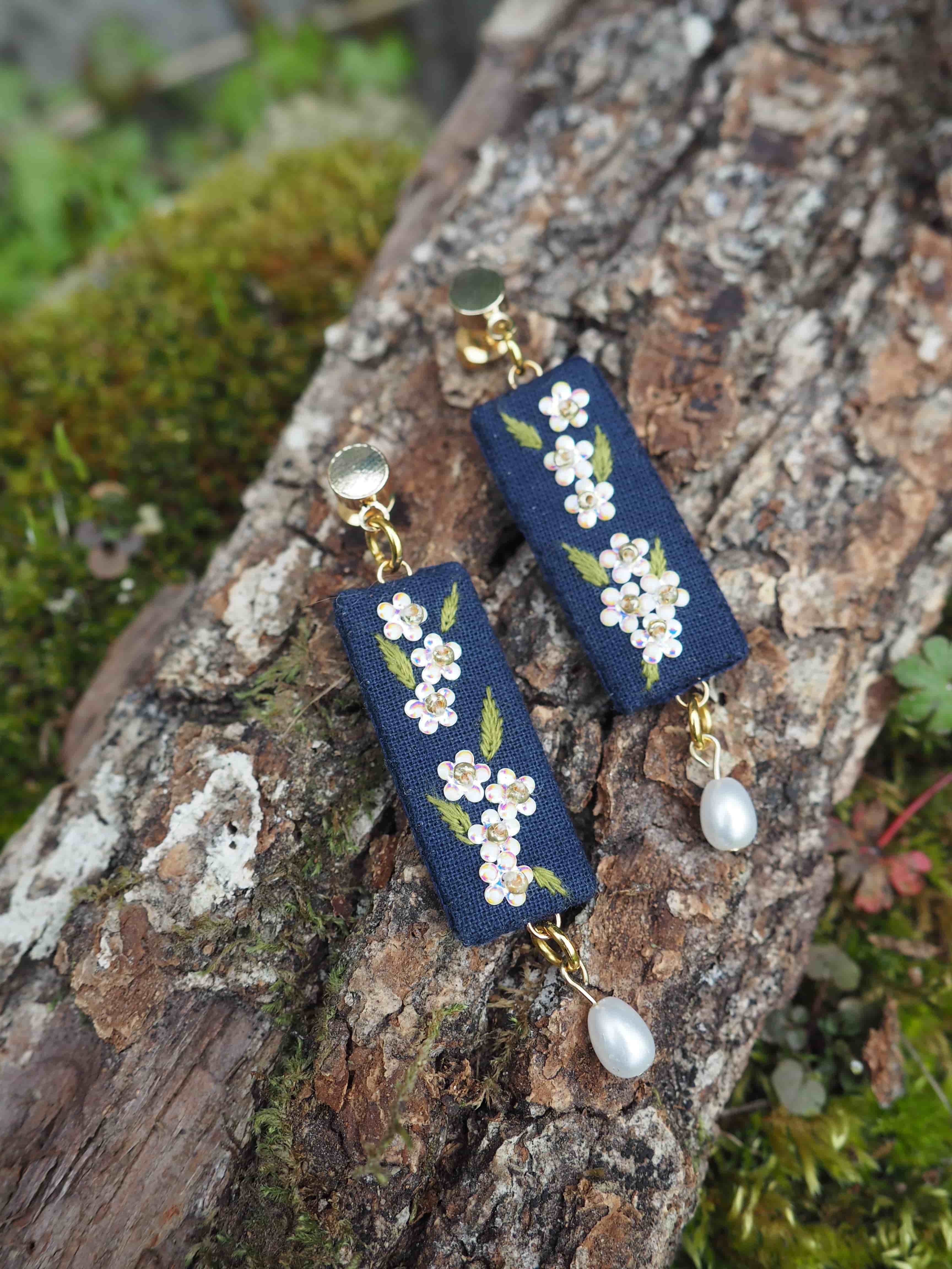 Ces boucles d'oreilles sont très élégantes et délicates. Elles sont composées d'une base en tissu bleu marine sur laquelle sont brodées de petites fleurs blanches avec des feuilles vertes. Au bas de chaque boucle d'oreille pend une perle blanche. Les attaches semblent être en métal doré. Les boucles d'oreilles sont photographiées sur un morceau d'écorce d'arbre recouvert de mousse, ce qui crée un beau contraste naturel avec les bijoux. Le style est à la fois romantique et artisanal, rappelant la broderie traditionnelle japonaise ou le style chinois.