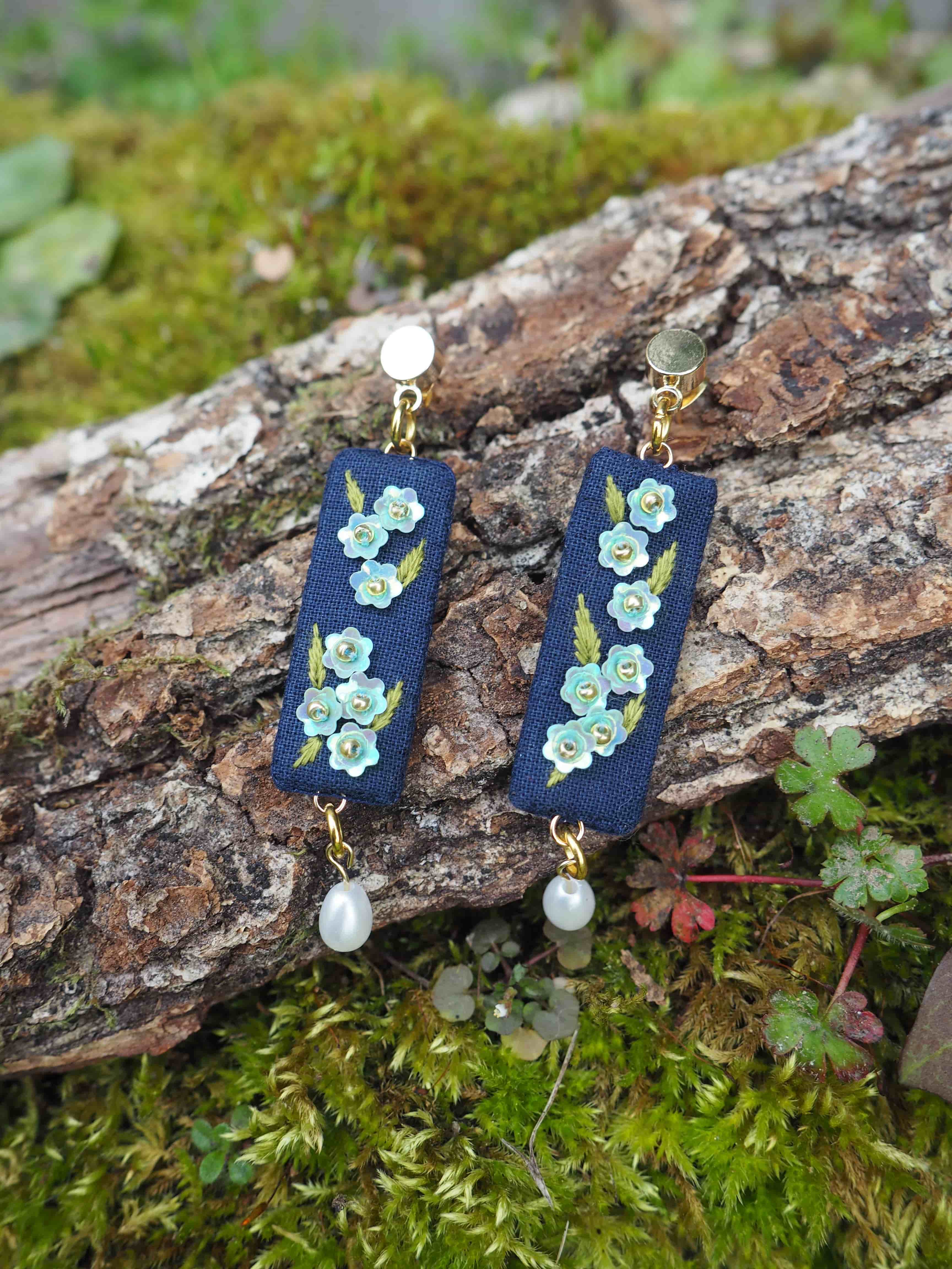 Cette image montre une paire de boucles d'oreilles artisanales posées sur un morceau d'écorce couvert de mousse. Les boucles d'oreilles sont composées de rectangles en tissu bleu marine brodés de petites fleurs bleues et vertes. Chaque boucle d'oreille est ornée d'une perle blanche nacrée en haut et en bas, reliées par des attaches dorées. Le cadre naturel avec la mousse verte et l'écorce texturée met joliment en valeur ces bijoux délicats et romantiques. Le style de ces boucles d'oreilles évoque un travail minutieux de broderie florale sur textile.