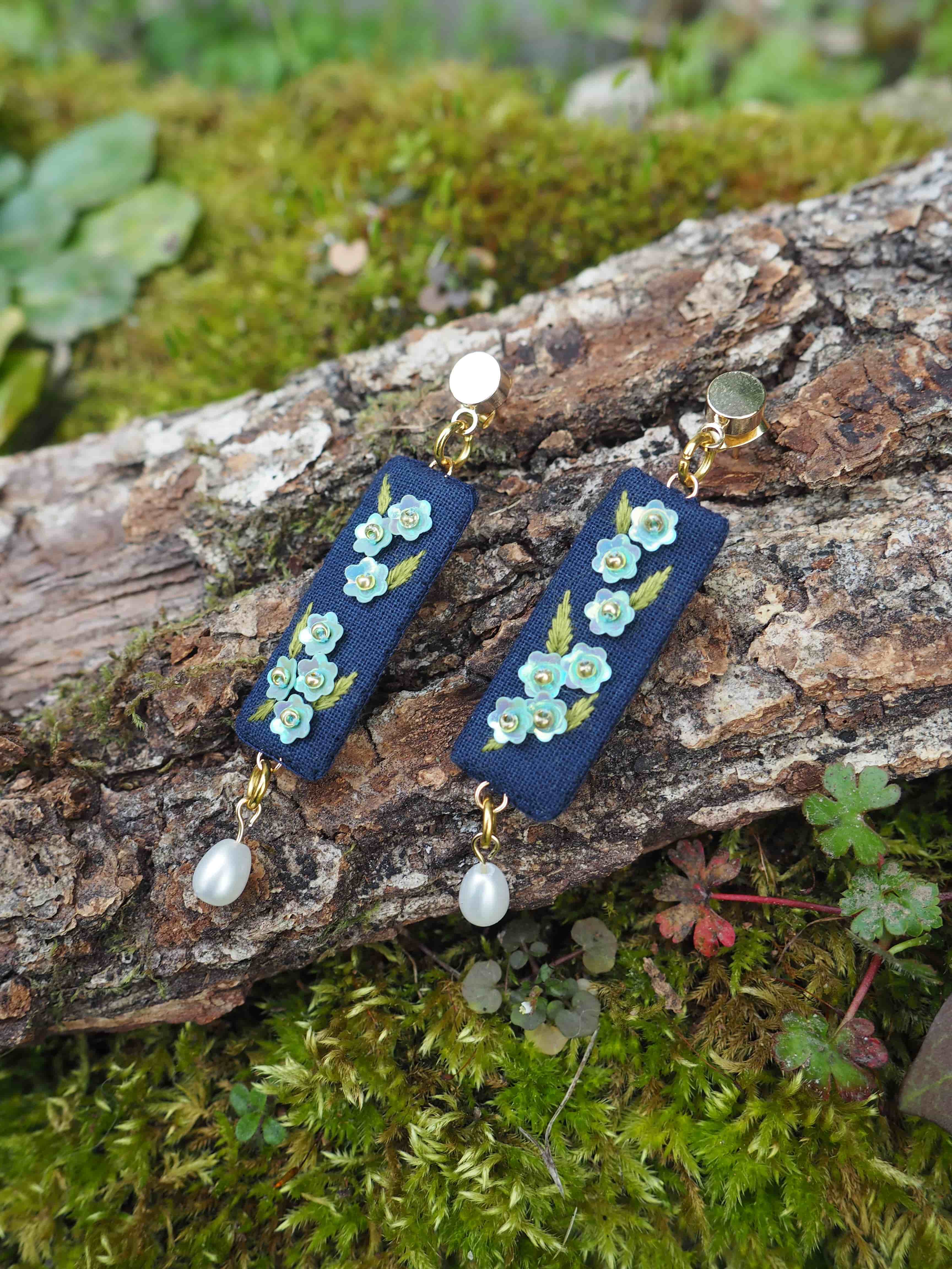 Cette image montre une paire de boucles d'oreilles artisanales posées sur un tronc d'arbre couvert de mousse. Les boucles d'oreilles sont composées d'une base en tissu ou cuir bleu marine, décorée de petites fleurs turquoise ou vert d'eau. Au bas de chaque boucle d'oreille pend une perle blanche nacrée. Les bijoux sont photographiés dans un cadre naturel, avec de la mousse verte et des petites plantes en arrière-plan, ce qui crée une ambiance naturelle et romantique. Le contraste entre le bleu des boucles d'oreilles et le vert de la mousse met particulièrement en valeur ces bijoux délicats.