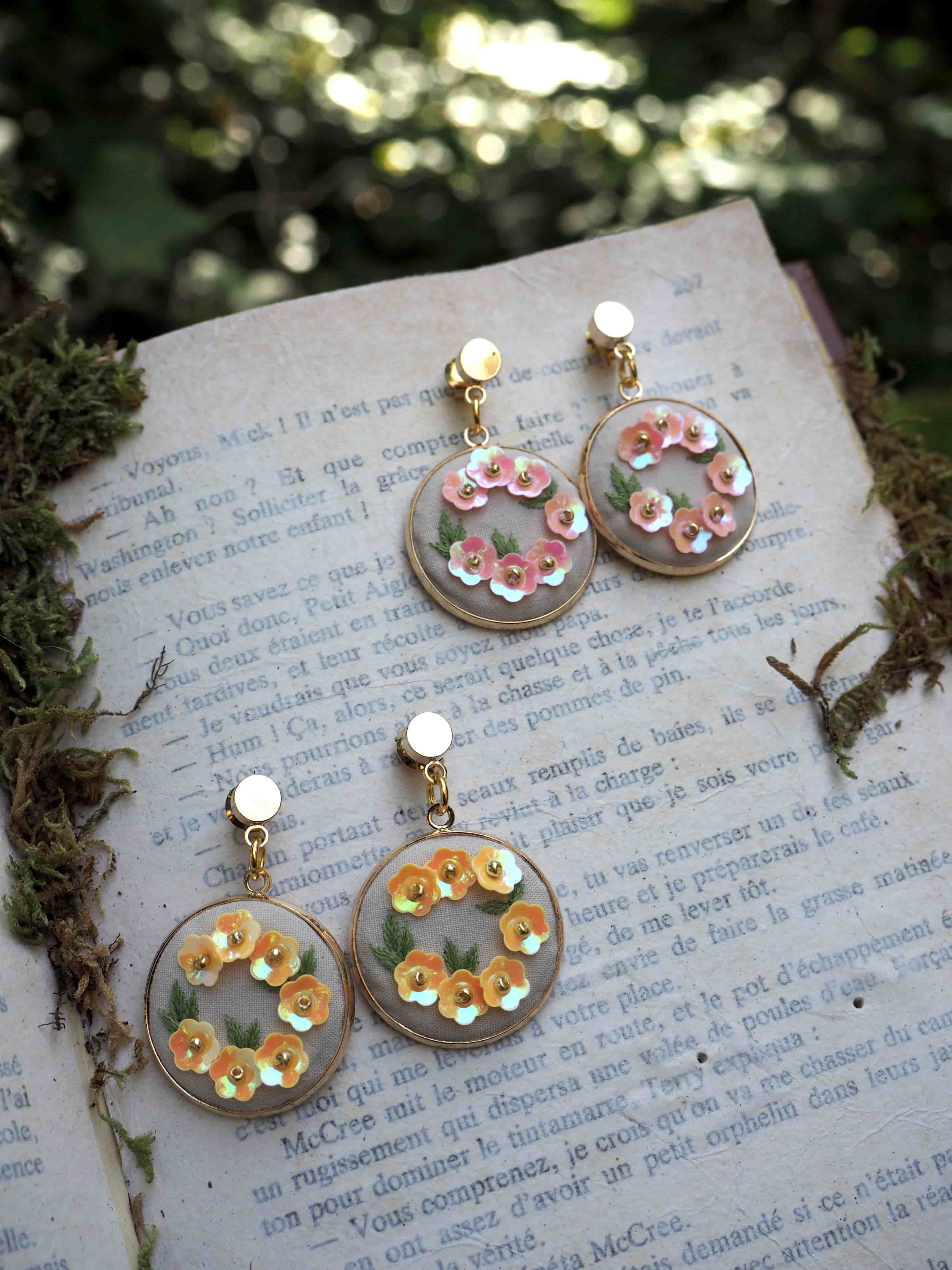 This image shows two pairs of delicate handmade earrings displayed on what appears to be an old French text or book page. The earrings feature a circular design with small decorative elements arranged in a ring pattern. One pair has pink/coral colored details while the other pair has yellow/gold colored accents. The earrings appear to be made with clear resin or acrylic bases with clip-on or post backings. They're photographed in natural lighting with some moss or greenery visible around the edges of the book page, creating a whimsical, vintage-inspired aesthetic. The overall composition has a romantic, artisanal quality that suggests these are handcrafted jewelry pieces.