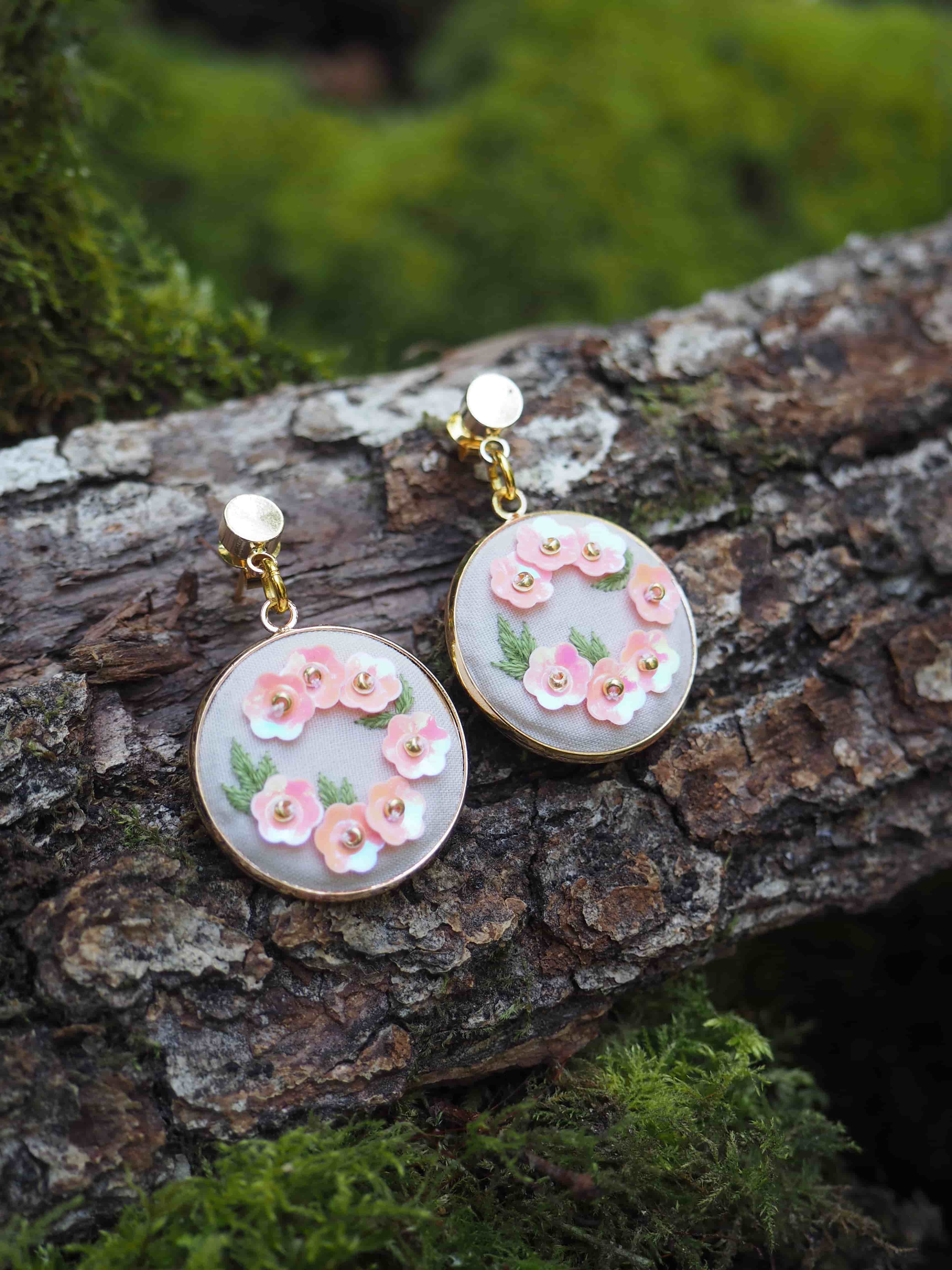 Cette image montre une paire de boucles d'oreilles artisanales délicates posées sur un morceau d'écorce d'arbre moussu. Les boucles d'oreilles sont de forme circulaire avec un fond blanc ou nacré, décorées de petites fleurs roses en relief et de feuilles vertes. Elles semblent être faites à la main, probablement en polymère ou en résine, avec des attaches dorées. La composition est très naturelle et romantique, avec le contraste entre la texture rugueuse de l'écorce et la délicatesse des bijoux. La mousse verte en arrière-plan ajoute une touche naturelle et féerique à la photo.
