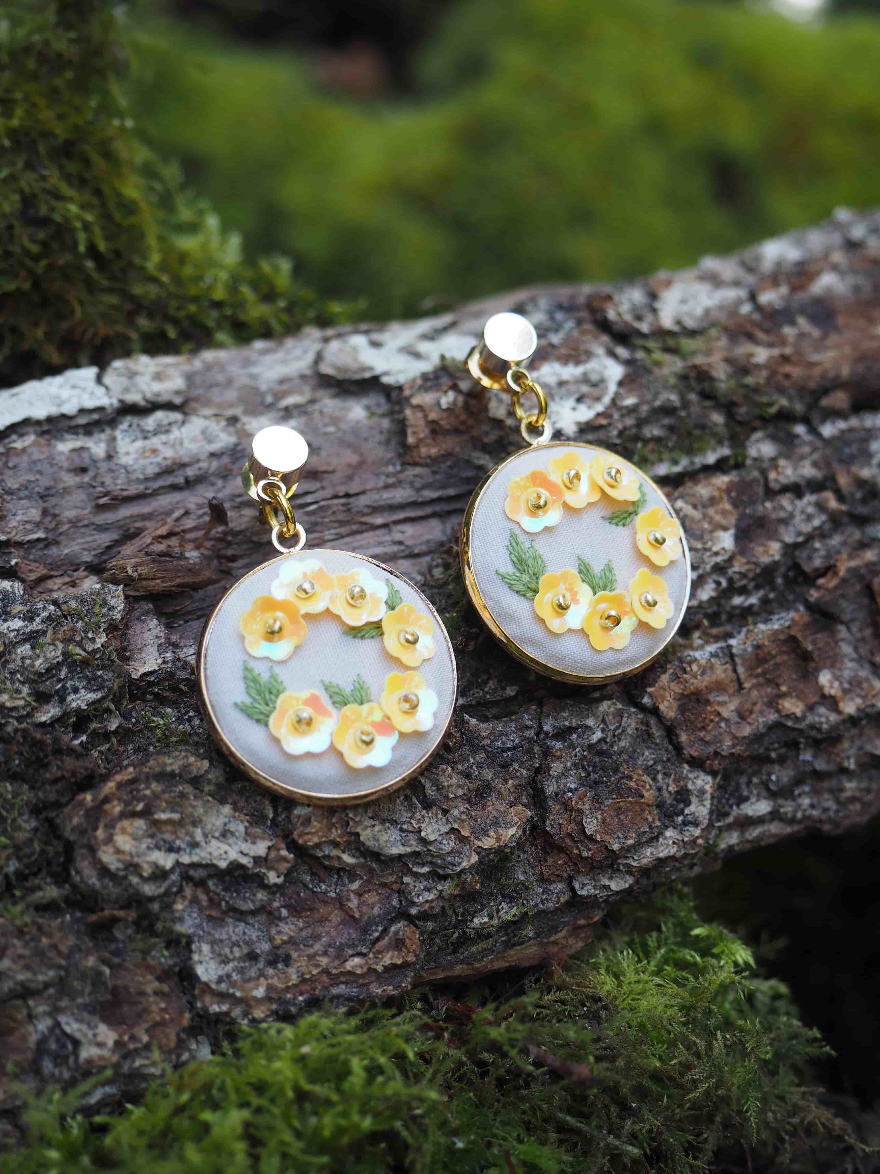 Cette image montre une paire de boucles d'oreilles artisanales délicates posées sur un morceau d'écorce d'arbre moussue. Les boucles d'oreilles sont de forme circulaire avec un fond blanc nacré, décorées de petites fleurs jaunes et de feuilles vertes peintes à la main. Elles semblent être faites dans un style vintage ou romantique. Les pendentifs sont attachés à des tiges dorées. La photo est prise dans un environnement naturel, avec de la mousse verte floue en arrière-plan, ce qui crée un beau contraste avec les bijoux délicats. L'éclairage naturel met en valeur les détails des motifs floraux sur les boucles d'oreilles.