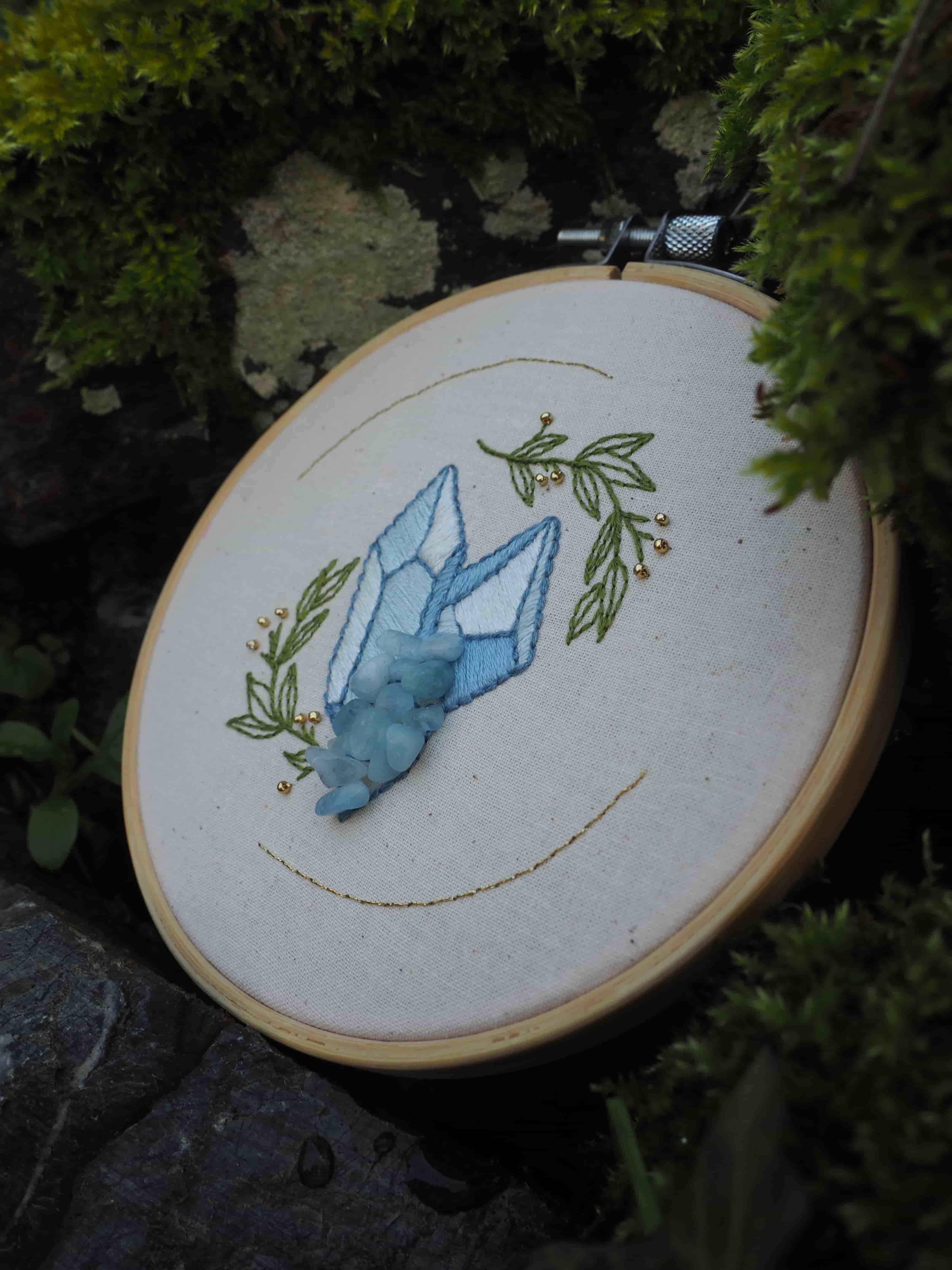 Cette image montre un ouvrage de broderie dans un cercle à broder traditionnel en bois. La broderie représente un cristal ou une pierre précieuse de couleur bleu clair, entourée d'éléments végétaux brodés en vert olive. Le motif est réalisé sur un tissu blanc, probablement de la toile à broder. La photo est prise dans un environnement extérieur, avec de la verdure floue en arrière-plan, ce qui crée une belle mise en scène naturelle pour cette pièce d'artisanat. Le travail de broderie semble délicat et soigné, avec des points qui créent un bel effet d'ombrage sur le cristal.