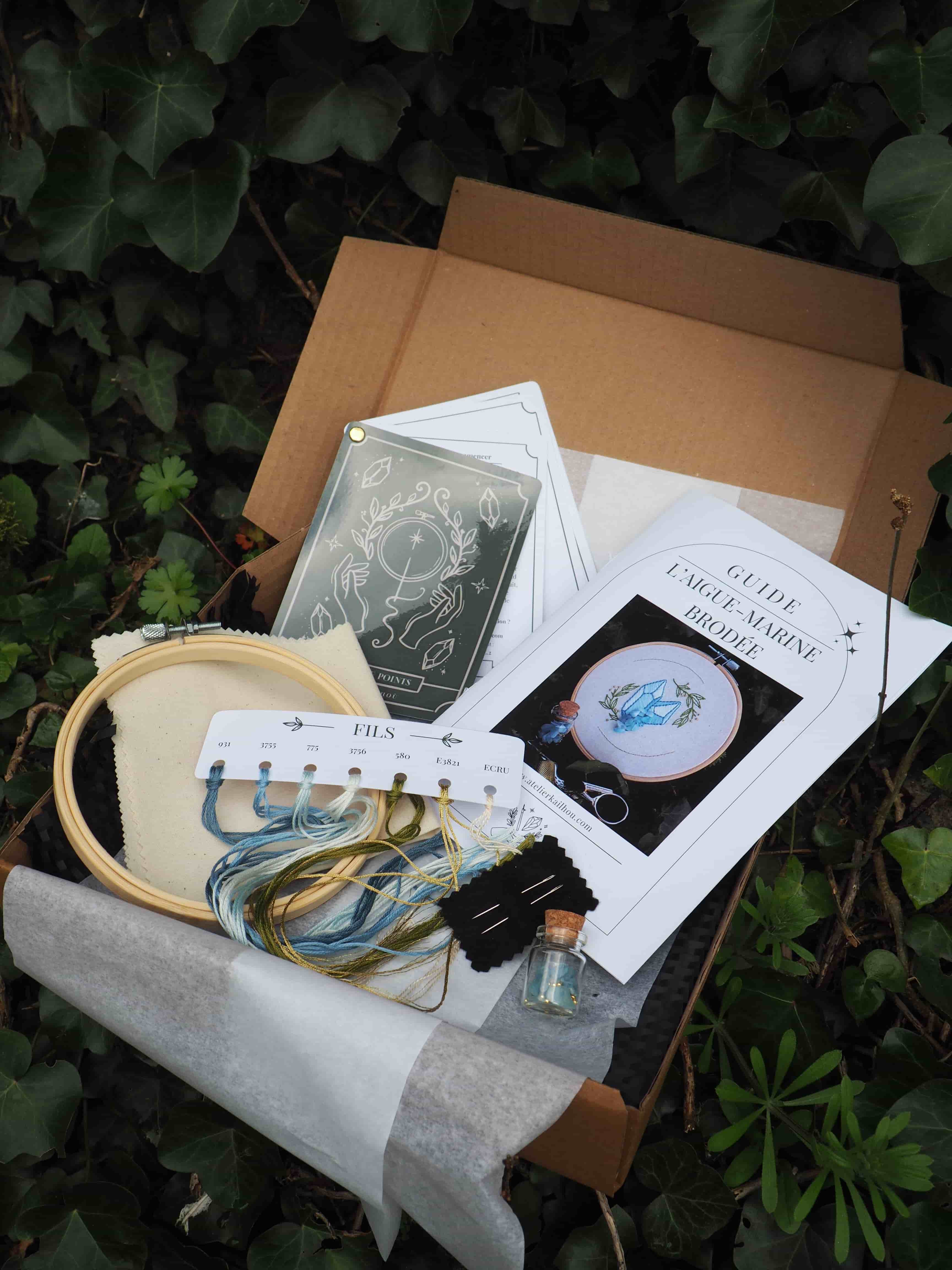 Cette image montre ce qui semble être un kit de broderie déballé. On peut voir : - Un cercle à broder en bois
- Des fils de broderie dans des tons de bleu et vert
- Des instructions ou patrons sur papier
- Une boîte en carton marron qui contenait probablement le kit
- Un fond de feuillage vert sombre
- Ce qui semble être du tissu blanc ou gris clair C'est typique d'un kit de broderie "prêt à commencer" qui contient tous les éléments nécessaires pour réaliser un projet de broderie. L'image a une ambiance artisanale et créative, suggérant un nouveau projet artistique sur le point de débuter.