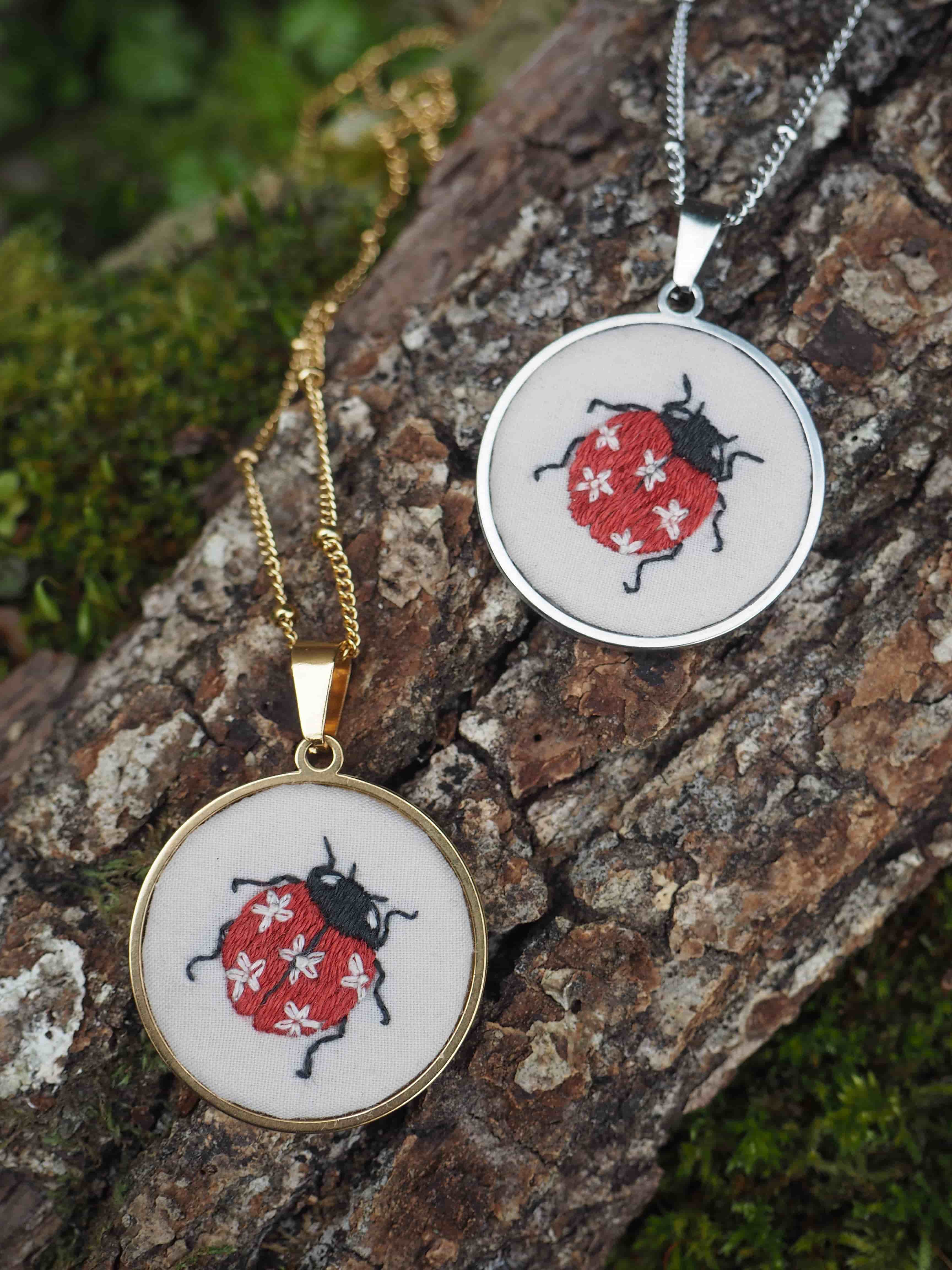 Cette image montre deux pendentifs similaires posés sur ce qui semble être de l'écorce d'arbre avec de la mousse. Les pendentifs représentent des coccinelles rouges avec des points blancs, brodées ou peintes sur un fond blanc. L'un des pendentifs est monté sur une chaîne dorée, tandis que l'autre est sur une chaîne argentée. Les pendentifs sont de style artisanal et délicat, avec un design simple mais charmant. La composition de la photo met en valeur le contraste entre les bijoux raffinés et la texture naturelle rugueuse de l'écorce.