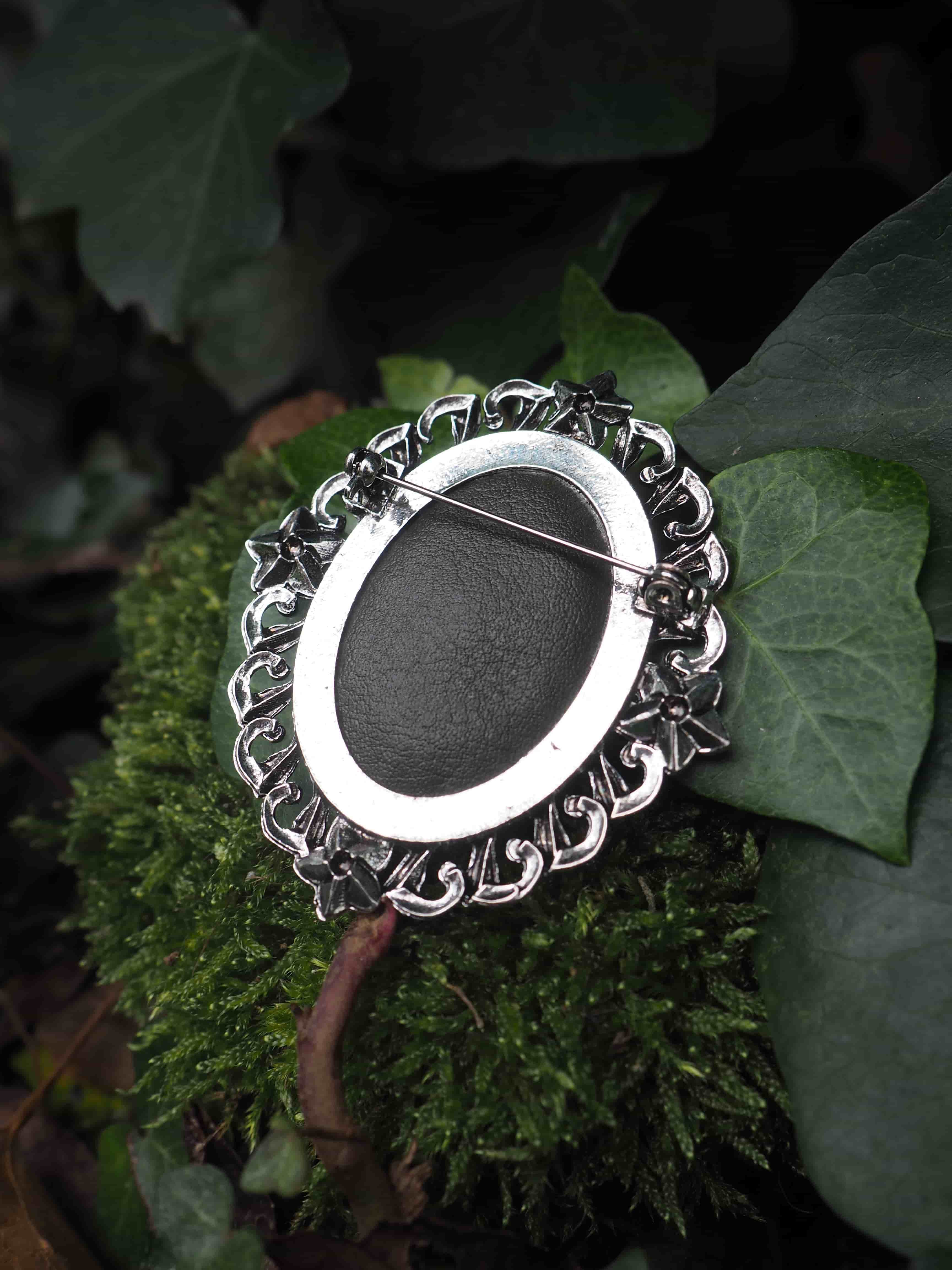 Cette image présente une broche ou un pendentif de style vintage posé délicatement sur des feuilles vertes dans un environnement naturel.  **Caractéristiques du bijou :**
- Forme ovale avec un centre noir mat (probablement en onyx ou une pierre similaire)
- Monture en métal argenté (vraisemblablement de l'argent) avec des motifs décoratifs ciselés
- Bordure ornementée de volutes et de motifs floraux en relief
- Style rétro ou victorien avec un travail artisanal détaillé **Mise en scène :**
- Le bijou repose sur une feuille verte texturée, possiblement une feuille de géranium ou de plante similaire
- L'arrière-plan montre d'autres feuillages dans des tons verts variés
- L'éclairage naturel met en valeur les contrastes entre le métal brillant, la pierre noire et la verdure environnante
- La composition crée une atmosphère élégante et naturelle, soulignant la beauté intemporelle du bijou Cette photographie met particulièrement bien en valeur le contraste entre l'objet manufacturé raffiné et son écrin végétal naturel.