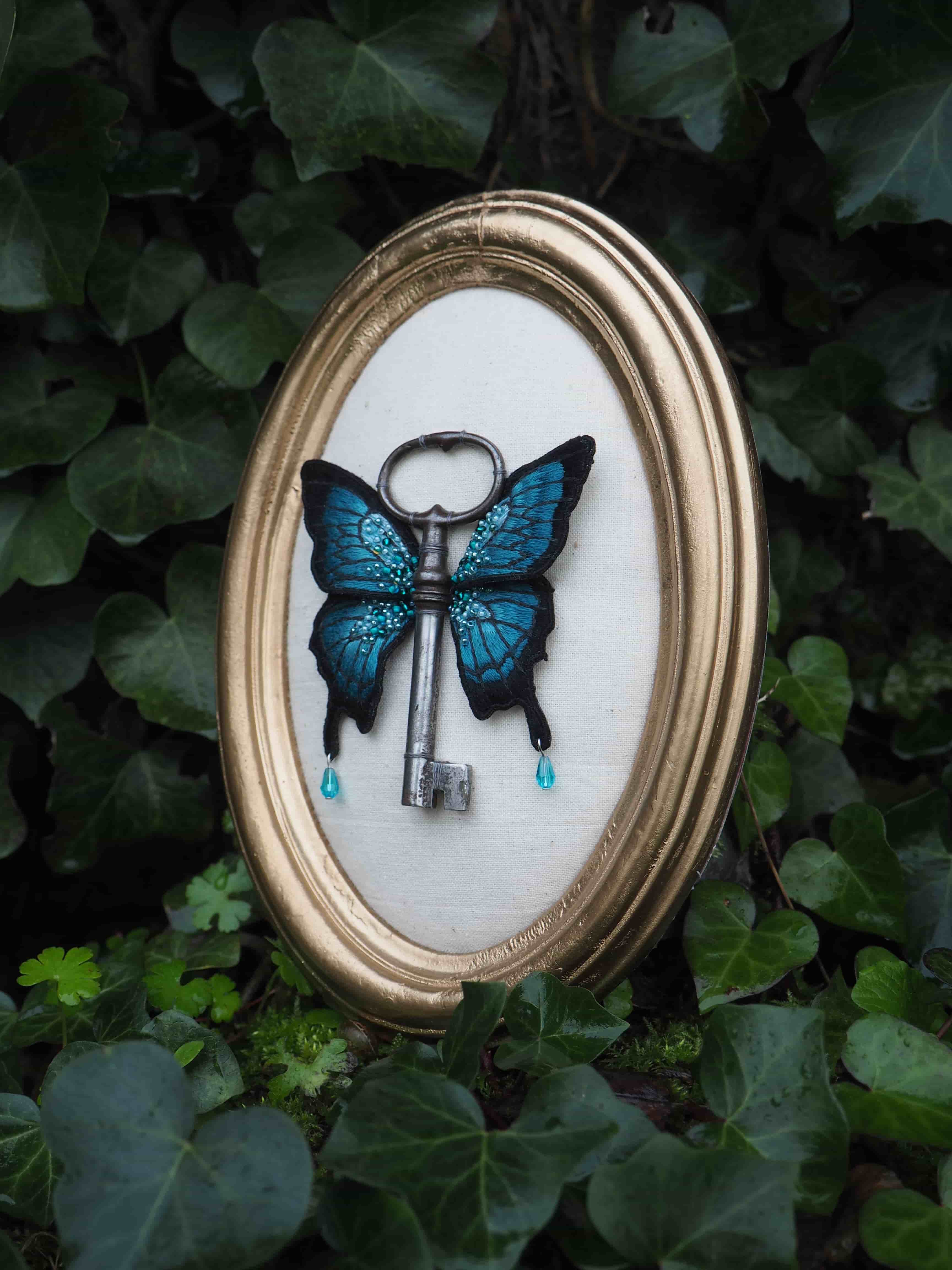 Cette image montre une création artistique encadrée très poétique : une clé ancienne transformée en papillon. La clé en métal est ornée d'ailes de papillon bleues turquoise, créant une pièce unique et fantaisiste. L'œuvre est présentée dans un cadre ovale doré sur un fond blanc, et l'ensemble est photographié au milieu de feuilles de lierre vert. Deux petites perles ou gouttes bleues pendent sous les ailes, ajoutant une touche délicate à la composition. C'est une belle fusion entre un objet utilitaire et la nature, évoquant un univers féerique et mystérieux.