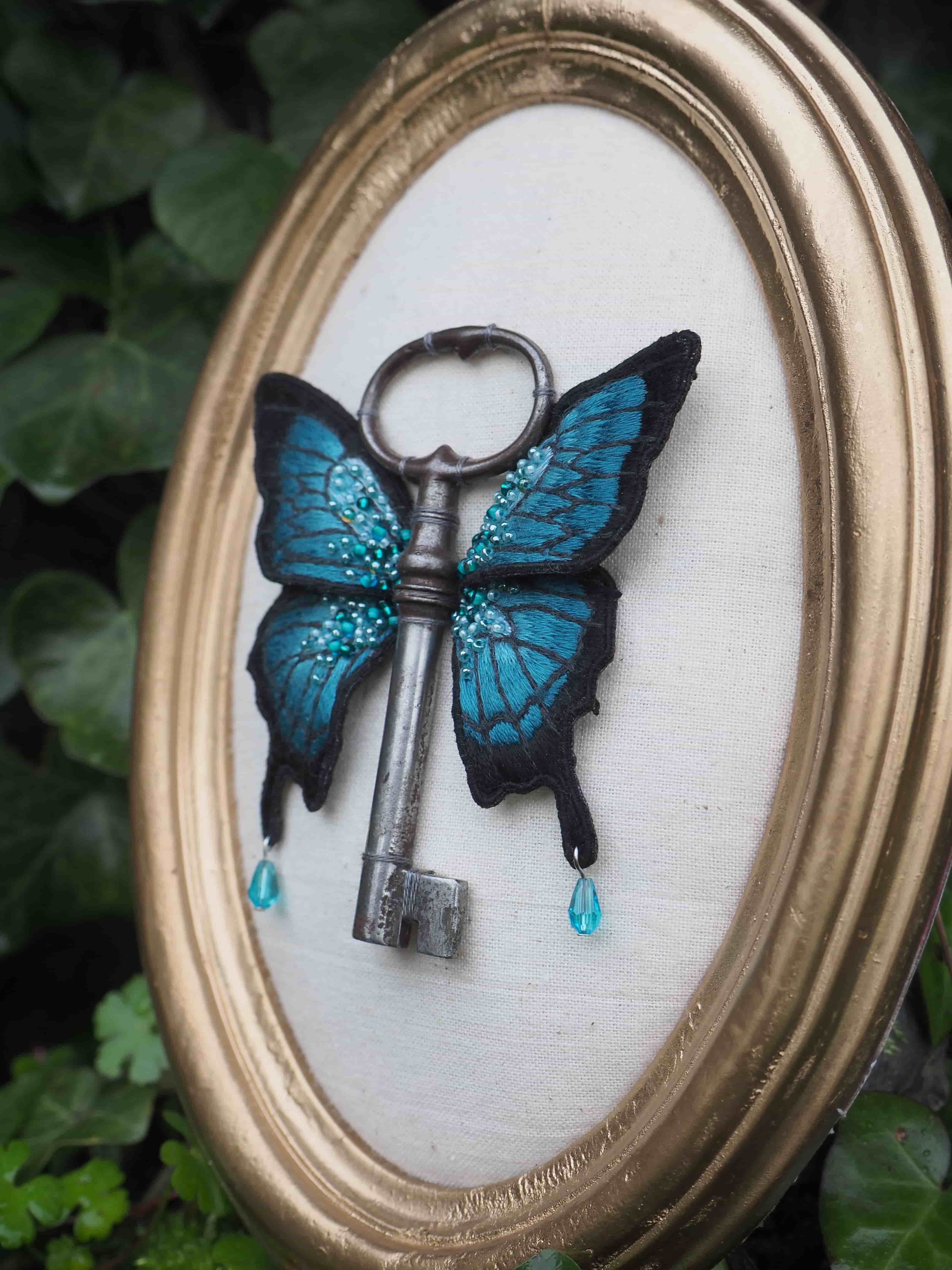 Cette image montre une création artistique très créative où une clé ancienne a été combinée avec des ailes de papillon bleues pour former un papillon-clé. L'œuvre est présentée dans un cadre ovale doré sur un fond blanc. Les ailes du papillon sont d'un bleu turquoise vibrant avec des motifs noirs, rappelant un morpho bleu. Deux petites gouttes bleues semblent tomber des ailes. La clé antique en métal sombre forme le corps du papillon. L'ensemble crée une pièce délicate et poétique qui mélange le dur et le délicat, l'ancien et le naturel. L'arrière-plan montre quelques feuilles vertes floues, suggérant que la photo a été prise dans un environnement naturel.
