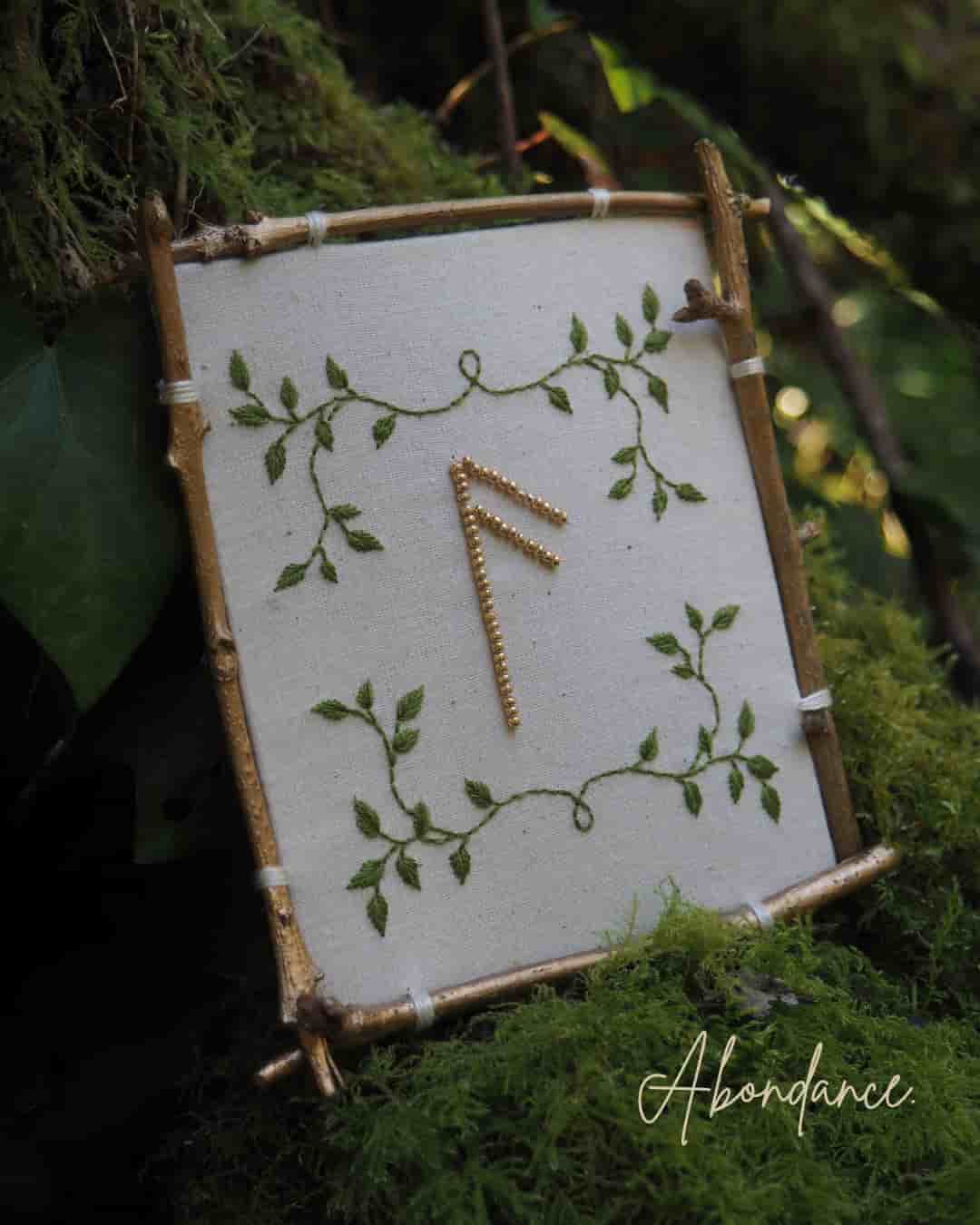 Cette image montre un symbole runique doré sur un fond blanc, encadré par un cadre rustique fait de petites branches ou de bois. Le symbole est décoré de motifs de feuilles vertes délicates qui s'entrelacent autour du cadre, créant un effet naturel et organique. Le mot "Abundance" (Abondance) est écrit en bas de l'image en écriture cursive élégante. L'ensemble est photographié sur un fond de mousse verte, donnant une ambiance très naturelle et mystique. Cette composition semble être inspirée par l'art païen ou la spiritualité nordique, où les runes sont souvent utilisées comme symboles de pouvoir ou de manifestation.