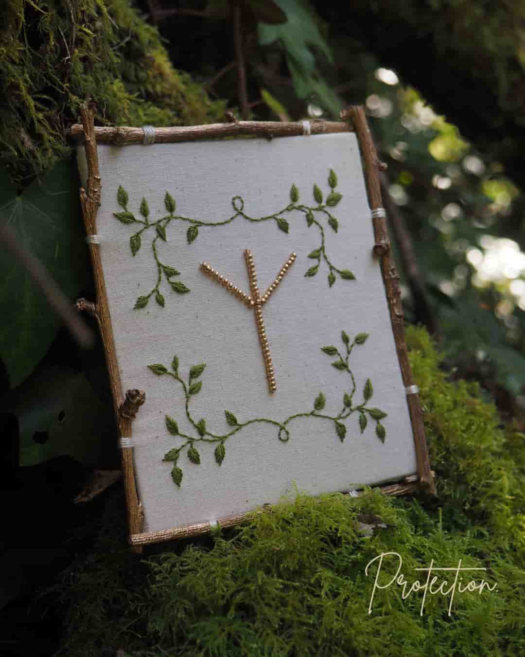 Cette image montre un symbole runique ou une rune Algiz (ᛉ) créée avec ce qui semble être des petites branches ou des brindilles, disposée sur un fond de tissu blanc ou écru. Le tissu est encadré par des branches naturelles formant un cadre rustique. Le fond est décoré d'un motif de feuilles vertes peintes ou brodées qui s'enroulent délicatement autour du symbole central. L'ensemble est photographié sur un lit de mousse verte, donnant une ambiance très naturelle et forestière. Le mot "Protection" apparaît en bas de l'image, ce qui est approprié car la rune Algiz est traditionnellement associée à la protection dans la tradition nordique. L'ensemble dégage une atmosphère païenne et naturelle très harmonieuse.